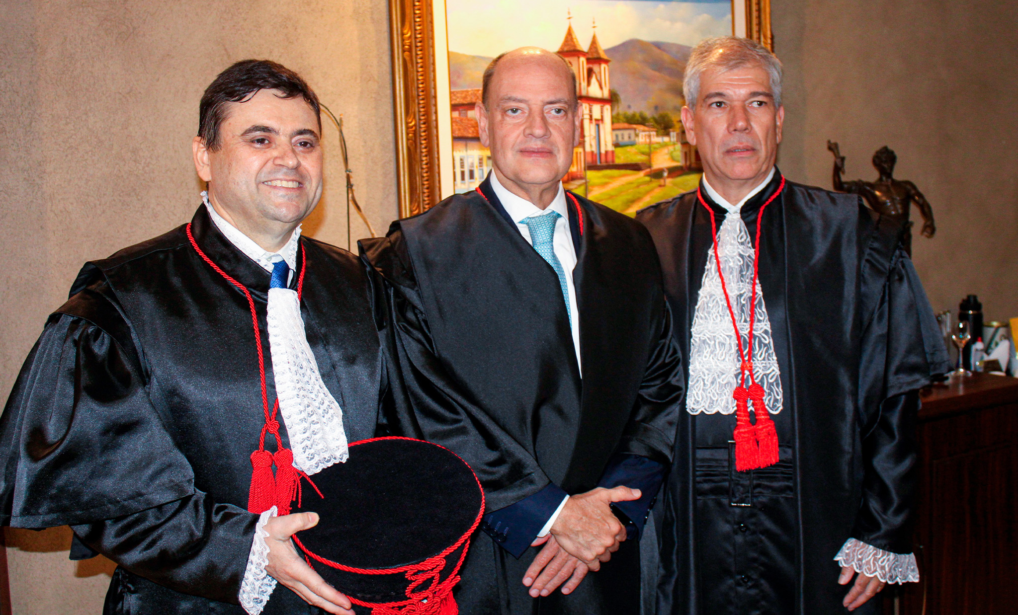 A imagem mostra três homens posando lado a lado em um ambiente interno, trajando vestes formais do Judiciário. Dois deles usam togas pretas com detalhes em renda branca e cordões vermelhos, típicas de magistrados, enquanto o homem ao centro veste terno escuro com gravata. O homem à esquerda segura um chapéu preto com detalhes vermelhos, parte do traje cerimonial. Os três têm expressão séria e postura formal, sugerindo um momento institucional importante, como uma cerimônia de posse. Ao fundo, há um quadro decorativo com paisagem e uma escultura, compondo um ambiente elegante e solene. 