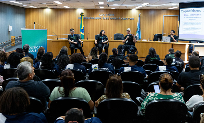 Imagem do auditório Desembargador José Navega Cretton. Ao fundo os convidados do evento falando para a plateia lotada de participantes. 