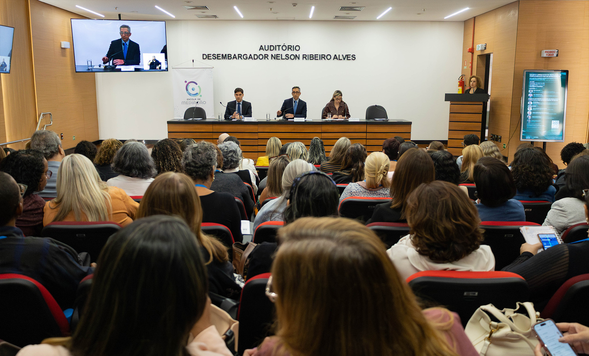 A imagem mostra um auditório lotado durante um seminário ou palestra promovida pela Escola de Mediação. O público ocupa todas as cadeiras do espaço e acompanha atentamente a mesa de debates posicionada à frente do auditório. No palco, três participantes estão sentados atrás de uma bancada. Ao centro, o desembargador Cesar Cury, que fala ao microfone enquanto os demais acompanham a apresentação. À direita da imagem, uma mulher está posicionada em um púlpito. Ao fundo, na parede branca, aparece a identificação do local: “Auditório Desembargador Nelson Ribeiro Alves”. Um telão suspenso exibe, em tempo real, a imagem ampliada do palestrante. O ambiente possui iluminação clara, acabamento em madeira e estrutura moderna.
