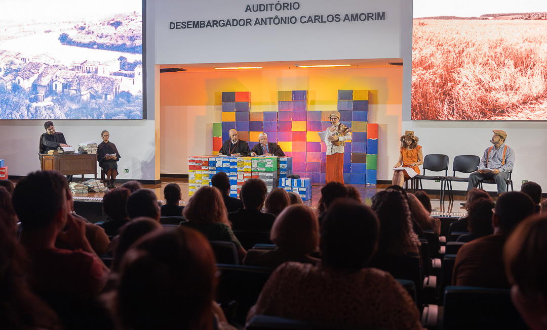 A imagem mostra o auditório Desembargador Antônio Carlos Amorim” com o público sentado e atento a apresentação no palco. No alto, ao centro, há a identificação do espaço: “Auditório Desembargador Antônio Carlos Amorim”. No palco, acontece uma encenação teatral. Os atores estão distribuídos em diferentes posições: à esquerda, dois personagens sentados atrás de uma mesa com uma grande quantidade de papéis empilhados, sugerindo um ambiente burocrático ou judicial. Ao centro, outros personagens também estão sentados atrás de uma mesa coberta por caixas coloridas empilhadas, formando um cenário geométrico e simbólico. À direita, há mais atores sentados e uma personagem em pé, lendo ou discursando a partir de um papel. O cenário combina elementos cênicos e visuais: ao fundo, painéis coloridos em formato de blocos criam um contraste com duas grandes telas laterais, que exibem imagens de paisagens rurais, como plantações e áreas do campo. 