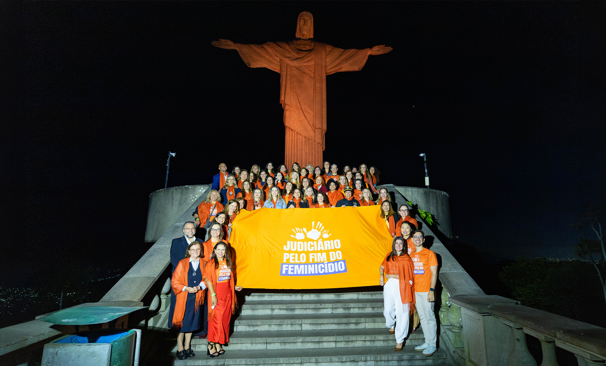 Fotografia noturna registrada na base do monumento ao Cristo Redentor, no Rio de Janeiro. Ao centro da imagem, dezenas de pessoas estão reunidas sobre a escadaria que leva ao monumento, posando para uma foto oficial. A maioria veste camisetas ou peças de roupa na cor laranja, em referência à campanha de conscientização pelo enfrentamento à violência contra a mulher. O grupo segura uma grande faixa laranja com a inscrição em branco e azul: "Judiciário pelo Fim do Feminicídio", acompanhada de um símbolo formado por mãos estilizadas. Em primeiro plano, algumas participantes estão posicionadas à frente da faixa, também vestidas em tons de laranja. Ao fundo, o Cristo Redentor aparece totalmente iluminado por luz alaranjada, com os braços abertos, destacando-se contra o céu escuro e reforçando a identidade visual da campanha. Nas laterais da escadaria, são visíveis os guarda-corpos de concreto e parte da vegetação do local. A iluminação direcionada destaca o grupo e o monumento, enquanto a paisagem da cidade permanece discretamente visível ao fundo.