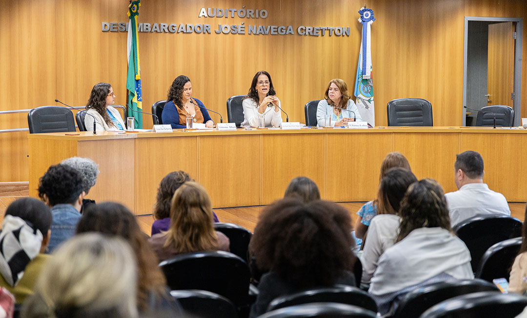 Imagem mostra quatro mulheres sentadas à mesa de palestra de um auditório com dezenas de pessoas sentadas à frente