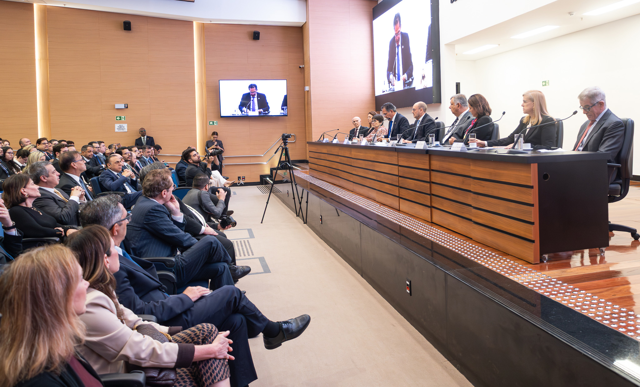 A imagem mostra um auditório durante um evento institucional ou jurídico, com grande público acompanhando uma mesa de autoridades posicionada sobre um palco. Ao fundo, um telão exibe a imagem ampliada de um dos participantes discursando ao microfone. Na mesa principal, composta por homens e mulheres trajando roupas sociais, os participantes estão sentados lado a lado diante de microfones, copos de água e documentos. À frente do palco, há uma câmera em tripé registrando a cerimônia. O público ocupa as cadeiras do auditório e acompanha atentamente a apresentação. O ambiente possui paredes em tons amadeirados, iluminação clara e estrutura moderna, reforçando o caráter formal do encontro.