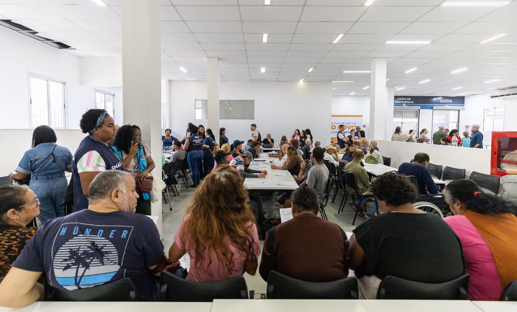 A imagem mostra um amplo salão de atendimento com grande circulação de pessoas. No centro do ambiente, diversas mesas estão ocupadas por usuários e profissionais, que conversam e realizam atendimentos. Em primeiro plano, várias pessoas estão sentadas em cadeiras, aguardando atendimento, enquanto outras permanecem em pé ao lado de uma coluna. Ao fundo, equipes identificadas por camisetas institucionais prestam orientações aos participantes. O espaço é bem iluminado, com paredes brancas, pé-direito alto e iluminação em LED. Também são visíveis placas de sinalização, como "Saída" e "Saída de Emergência", além de um banner institucional ao fundo e um extintor de incêndio instalado na parede à direita.