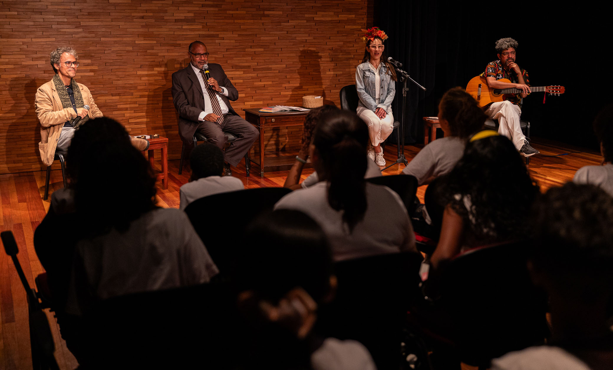 Foto mostra quatro pessoas santadas em cadeiras sobre um palco, à meia luz, sendo, da esquerda para a direita, um homem de cabelos claros  e casaco bege, em seguida outro, de terno escuro, óculos  falando ao microfone, ao seu lado, uma mulher de cabelos longos escuros com uma tiara de flores grandes na cabeça e vestindo cala branca e blusa jeans, e, por último um homem de camisa estampada, cabelos encaracolados, de pernas cruzadas e segurando um violão