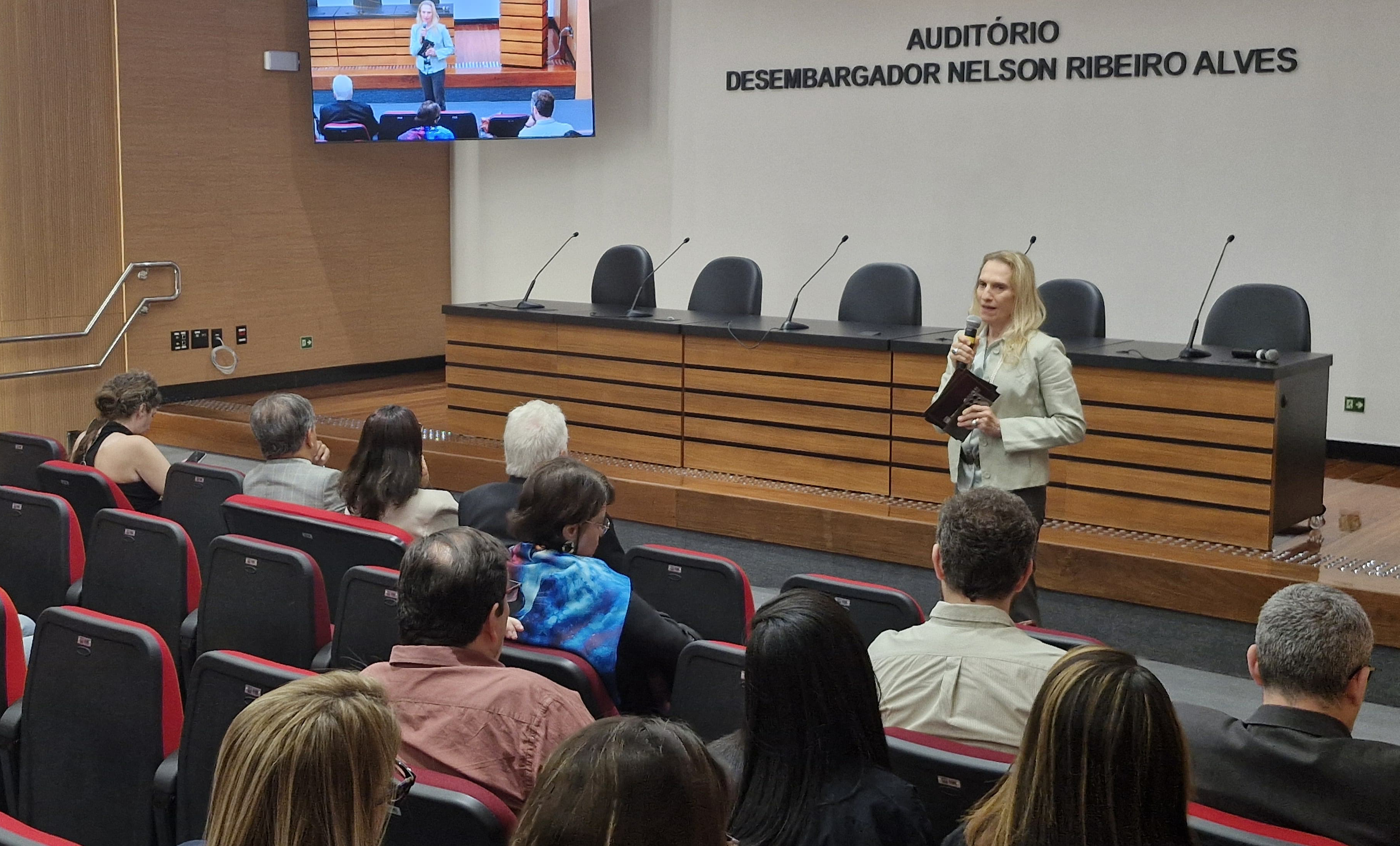 Foto mostra o ambiente de auditório, tendo uma mulher de cabelos loiros e casaco cinza falando ao microfone  para uma plateia sentada. A mulher está de pé e à frente da mesa principal do auditório, que também conta com telão ao fundo, à esquerda da tela