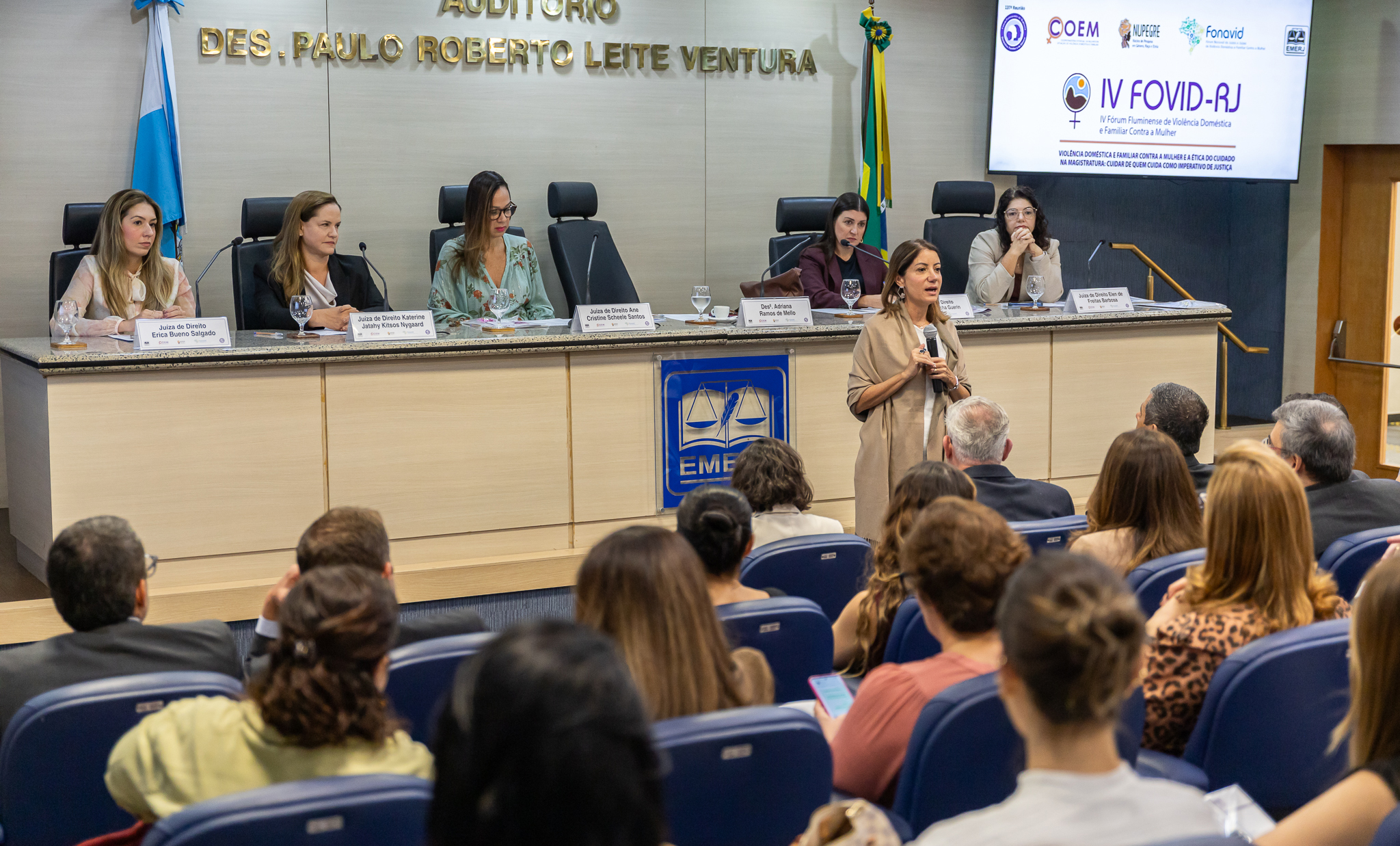 Em um auditório, uma mulher está em pé à frente da plateia, segurando um microfone e falando ao público. Ao fundo, seis mulheres compõem a mesa de autoridades, sentadas atrás de uma bancada equipada com microfones, copos de água e identificações nominais. Acima da mesa, há a inscrição "Auditório Des. Paulo Roberto Leite Ventura". À direita, um telão exibe a identidade visual do IV FOVID-RJ – IV Fórum Fluminense de Violência Doméstica e Familiar Contra a Mulher, com os logotipos das instituições organizadoras. As bandeiras do Brasil e do Estado do Rio de Janeiro estão posicionadas ao lado da mesa. Em primeiro plano, o público acompanha a apresentação, acomodado em poltronas azuis voltadas para o palco. O ambiente é iluminado por luz artificial e possui acabamento em madeira clara.