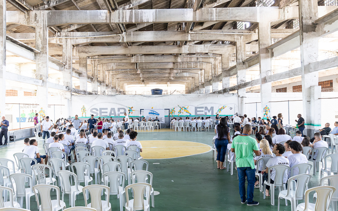 A imagem mostra um grande espaço coberto, semelhante a uma quadra poliesportiva, adaptado para a realização de um evento coletivo. O ambiente possui estrutura ampla, com vigas aparentes e iluminação natural entrando pelas laterais. No local, há diversas cadeiras plásticas brancas organizadas em fileiras, ocupadas por participantes que assistem a uma atividade central. Ao fundo, várias pessoas estão sentadas em mesas, indicando um momento de atendimento. Muitos dos presentes usam camisetas com a sigla “SEAP/RJ”. A quadra possui marcações esportivas no chão e paredes com pinturas e grafismos. 
