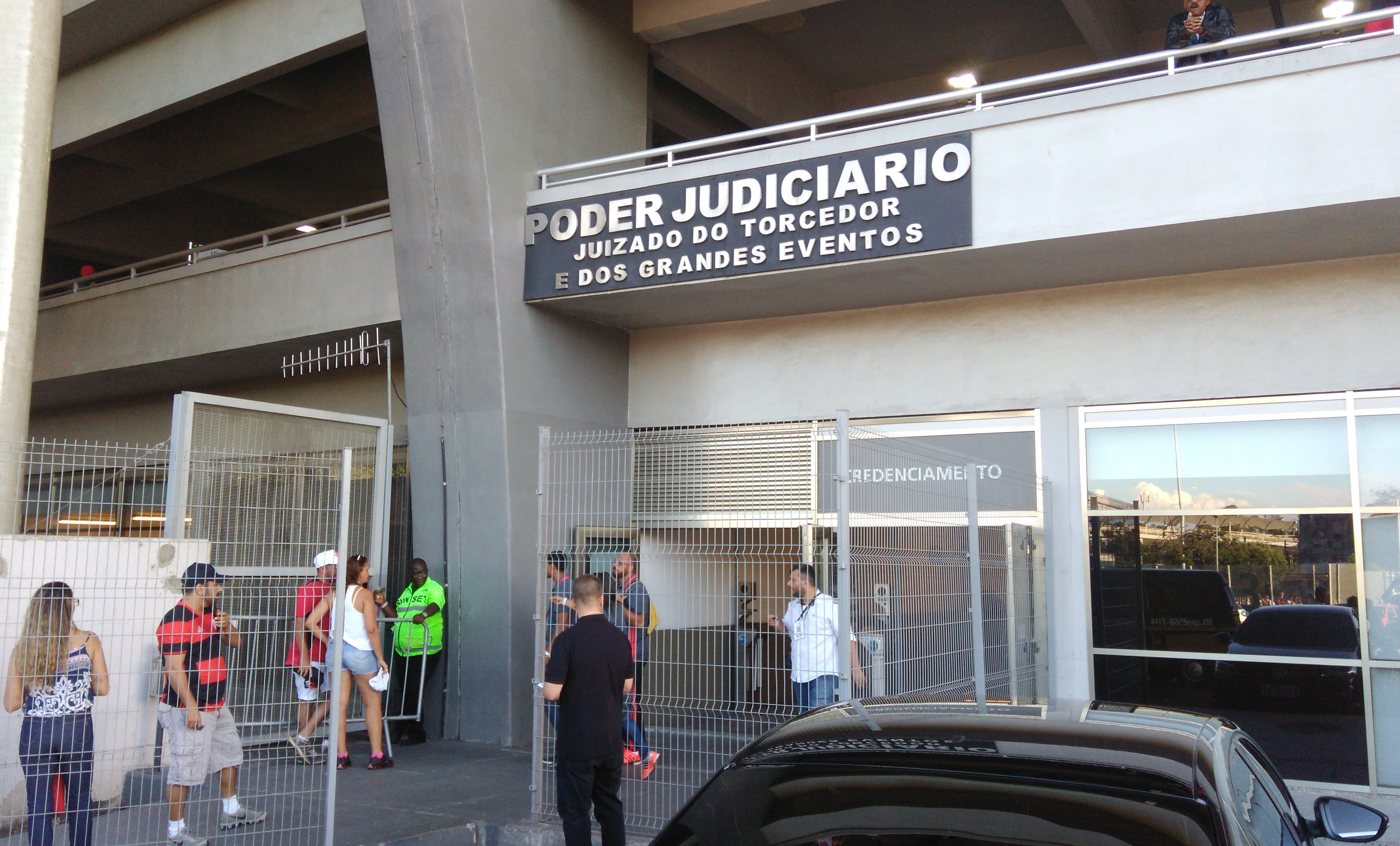 A imagem mostra a entrada do JUIZADO DO TORCEDOR E DOS GRANDES EVENTOS”, instalada na parte superior de uma estrutura de concreto do estádio do Maracanã. No nível térreo, há um portão metálico de grade que controla o acesso ao local. Algumas pessoas estão próximas à entrada, entre elas torcedores vestindo camisas de futebol, um agente de segurança com uniforme verde e outros indivíduos conversando. 