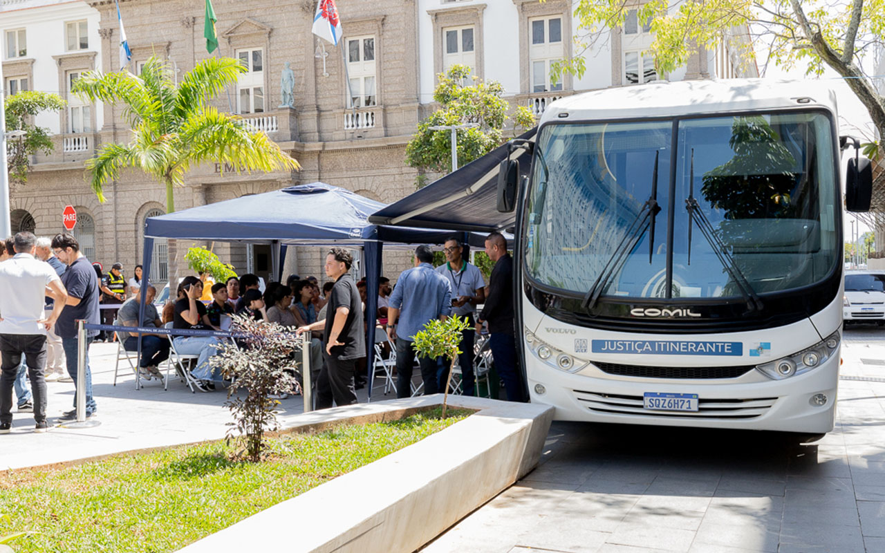 A imagem retrata uma ação de atendimento da Justiça Itinerante realizada ao ar livre, em frente ao prédio da Emerj. Em primeiro plano, à direita, há um ônibus branco identificado com a inscrição “Justiça Itinerante”, indicando que o veículo está sendo utilizado para levar serviços do Judiciário diretamente à população. Ao lado do ônibus, vê-se uma estrutura com tendas azuis e mesas, onde várias pessoas estão sentadas ou em pé, aguardando ou sendo atendidas. Há servidores e cidadãos interagindo, formando pequenas filas e grupos de conversa.