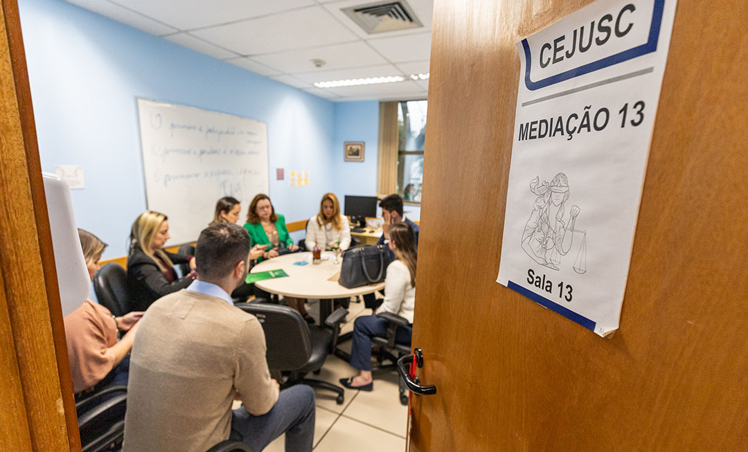 Imagem mostra pessoas ao redor de uma mesa em uma sala, em cena que demonstra uma negociação. Na porta da sala está escrito a palavra Cejusc - Mediação 13