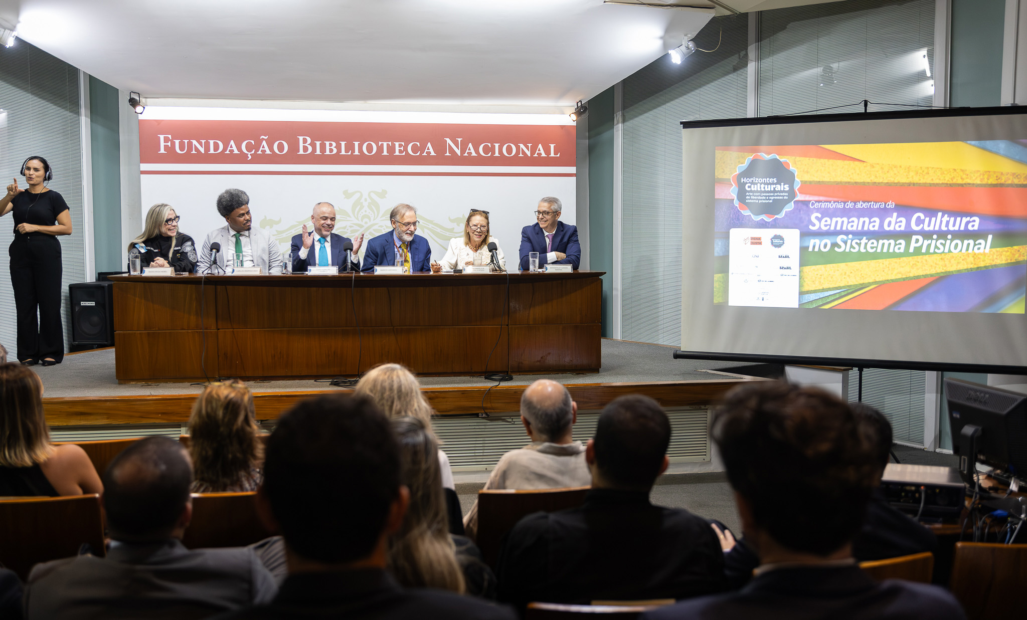 A imagem mostra uma mesa de autoridades em um evento institucional realizado em um auditório. Seis pessoas estão sentadas lado a lado atrás de uma bancada de madeira, com microfones e copos de água à frente, indicando que participam de uma mesa de abertura ou debate. São eles: coordenador da Senappen Carlos Rodrigo Dias; secretária da Seap Maria Rosa Nebel; desembargador do TJSP Luís Geraldo Lanfredi; presidente da Fundação Biblioteca Nacional, Marco Lucchesi; desembargadora Maria Angélica Guerra Guedes e Wanderley Sanan Dantas. Ao fundo, há um painel com a inscrição “Fundação Biblioteca Nacional”. À direita da imagem, um telão exibe uma apresentação com o título: “Cerimônia de abertura da Semana da Cultura no Sistema Prisional”, acompanhado de elementos gráficos coloridos e logotipos. Os participantes vestem trajes formais, como ternos e blazer, e demonstram postura atenta. 