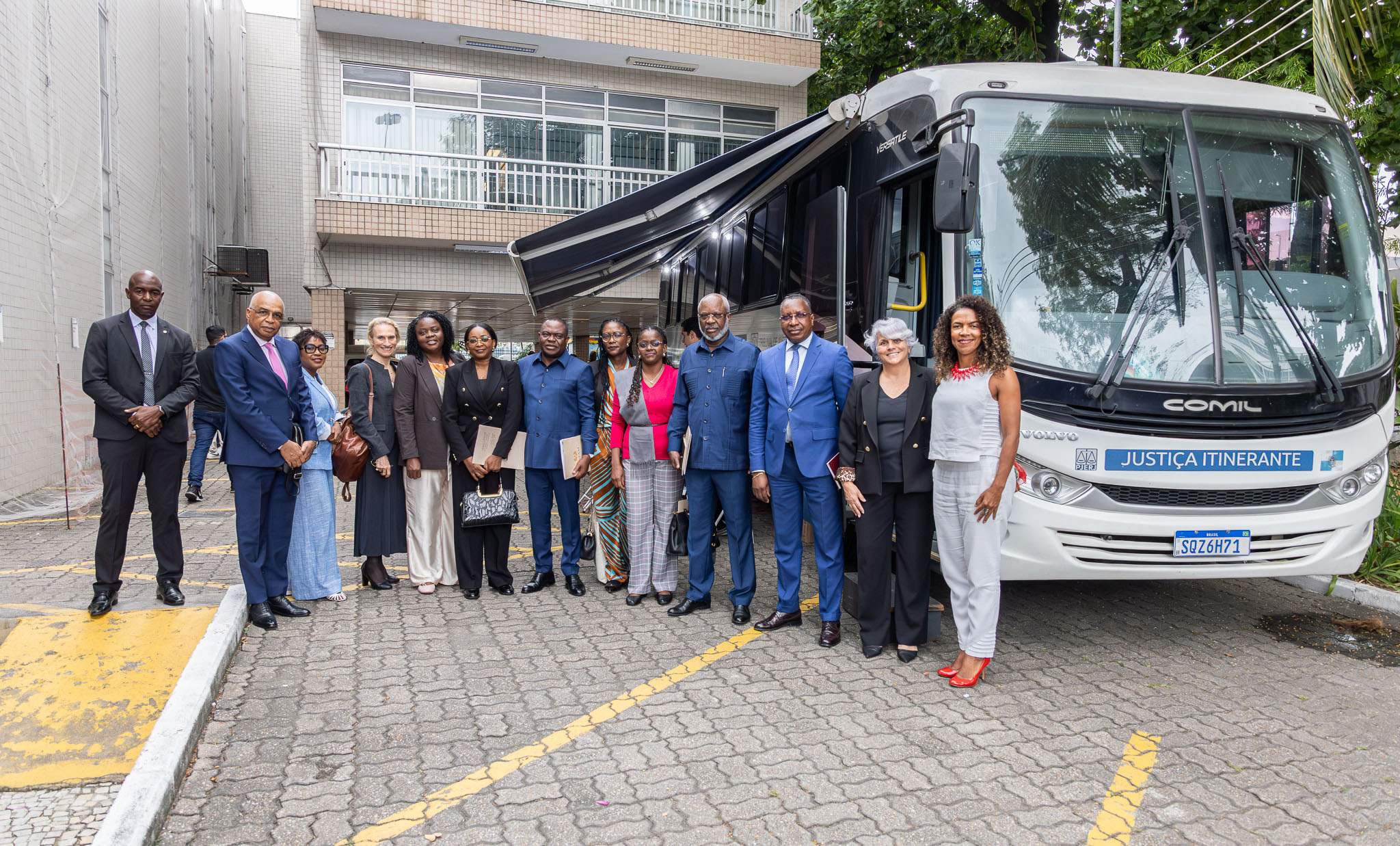 Imagem mostra um grupo de onze pessoas posando ao lado de um ônibus da Justiça Itinerante estacionado em uma área externa, em frente a um edifício. Os participantes estão alinhados diante do veículo, vestidos majoritariamente com roupas sociais em tons escuros e azuis. O ônibus branco possui a identificação “Justiça Itinerante” na parte frontal e um toldo lateral aberto. Ao fundo, aparecem árvores e a fachada do prédio com varandas e janelas amplas. O ambiente é iluminado pela luz do dia.