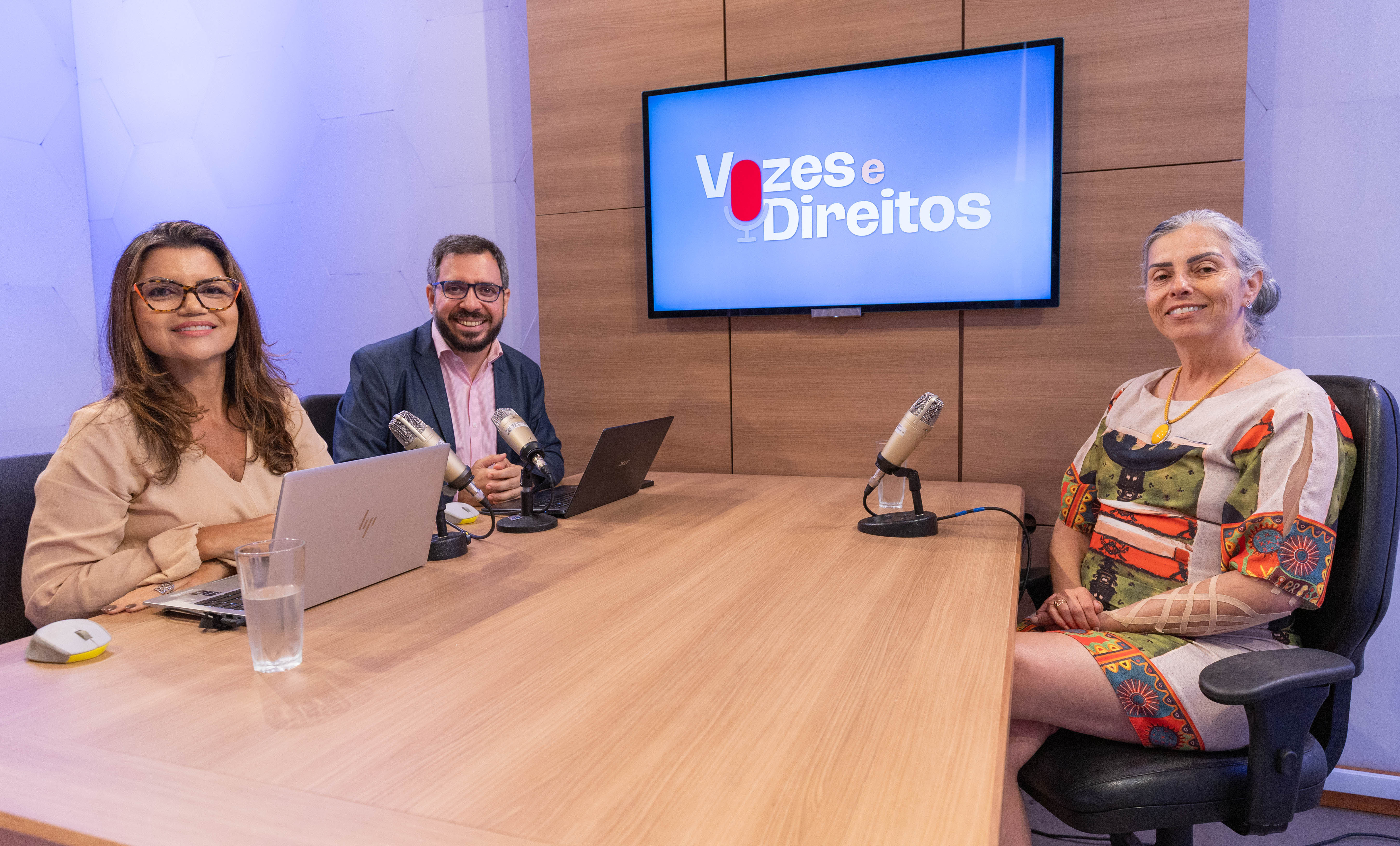 A imagem mostra um estúdio de gravação de podcast com três pessoas sentadas ao redor de uma mesa de madeira clara. À esquerda, uma mulher de cabelos castanhos e óculos sorri para a câmera, com um notebook aberto à sua frente e um microfone de mesa posicionado próximo a ela. Ao centro, um homem de barba, óculos e paletó escuro também sorri, sentado diante de outro notebook e de um microfone. À direita, uma mulher de cabelos grisalhos presos para trás, usando um vestido estampado colorido, está sentada em uma cadeira giratória e sorri em direção à câmera, com um microfone à sua frente. Na parede ao fundo, um monitor exibe a identidade visual do programa “Vozes e Direitos”, com o nome em letras brancas e um ícone de microfone vermelho sobre fundo azul. O ambiente tem revestimento de madeira e iluminação suave em tons azulados.