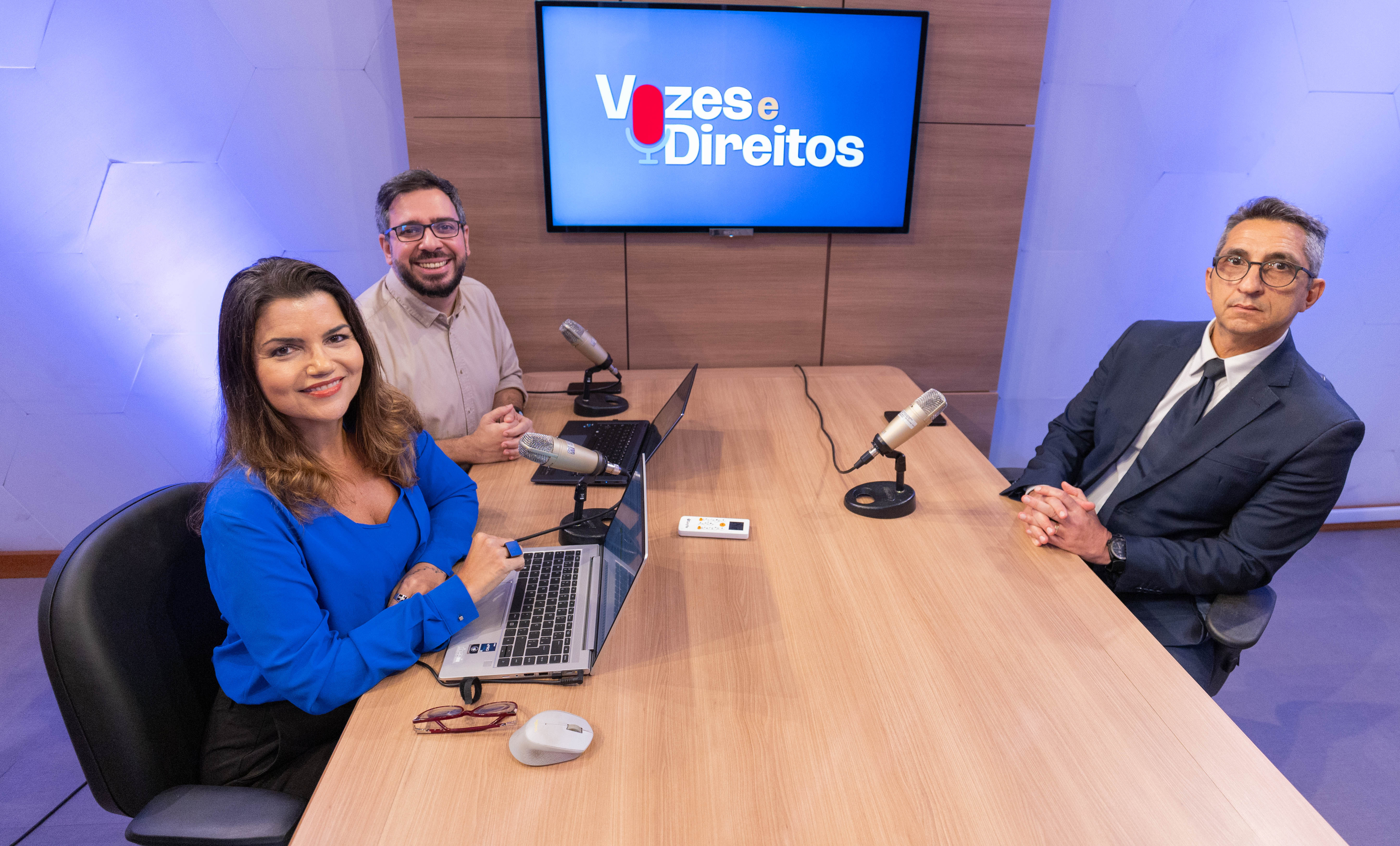 A imagem mostra um estúdio de gravação com três pessoas sentadas ao redor de uma mesa retangular de madeira. Sobre a mesa, há microfones de estúdio, notebooks abertos e um gravador de áudio.Ao fundo, um monitor exibe o nome “Vozes e Direitos”. À esquerda, uma mulher veste blusa azul, está sentada em frente a um notebook e sorri para a câmera. No centro, um homem com barba e óculos também sorri, posicionado atrás de um microfone e de um computador portátil. À direita está o desembargador Cesar Cury Nupemec, de terno e gravata. 