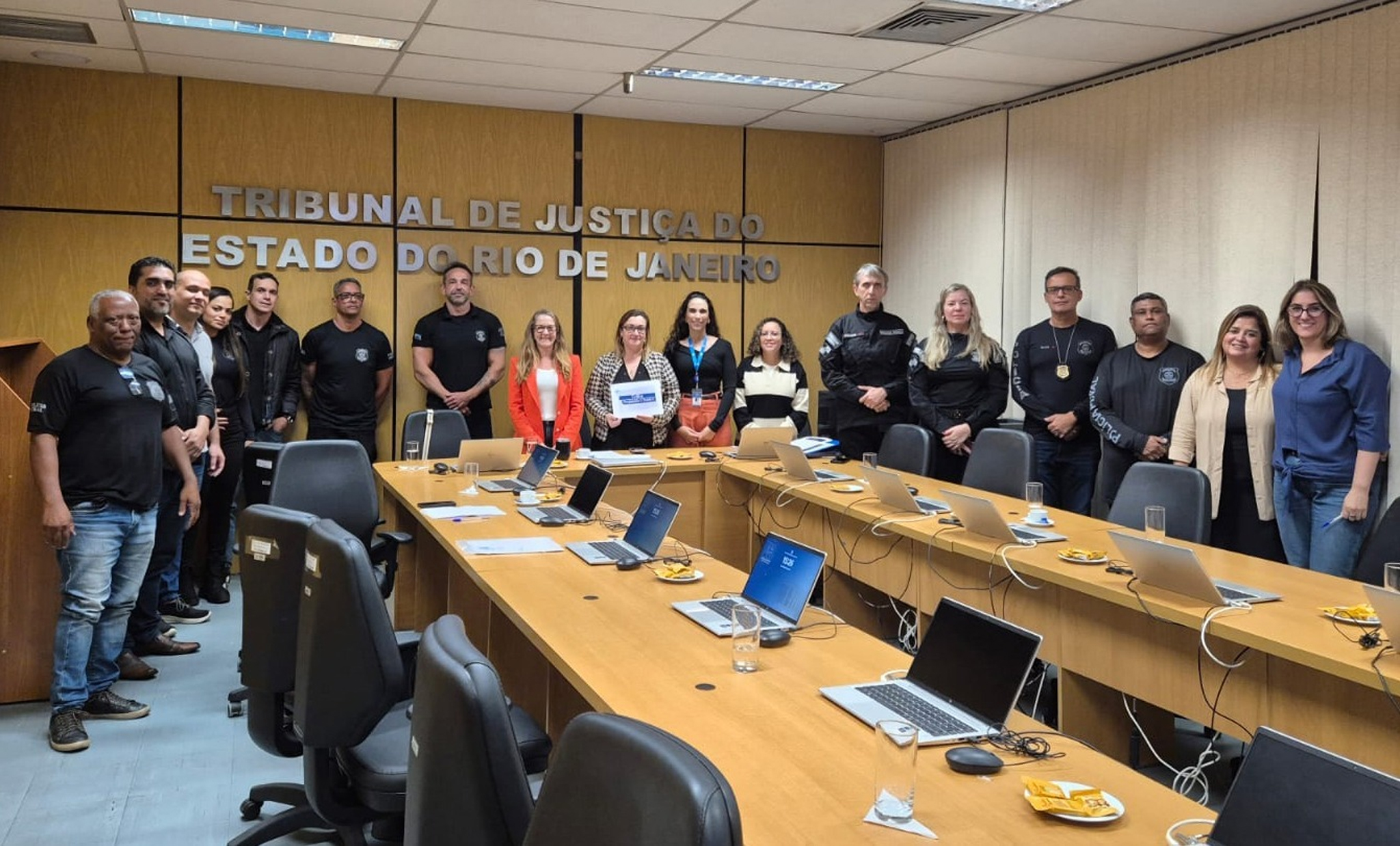 A imagem mostra um grupo de pessoas reunidas em uma sala de reuniões do Tribunal de Justiça do Rio de Janeiro (TJRJ). Os participantes estão posicionados lado a lado ao fundo do ambiente, voltados para a câmera, em um registro institucional. A sala possui paredes revestidas em madeira clara e, ao centro da parede principal, destaca-se a inscrição “Tribunal de Justiça do Estado do Rio de Janeiro” em letras metálicas. À frente do grupo, há uma grande mesa de reuniões em formato retangular, equipada com notebooks, copos de água, documentos e materiais de trabalho, sugerindo a realização de um encontro ou atividade de capacitação. Entre os participantes, há homens e mulheres com diferentes perfis profissionais. Alguns vestem trajes sociais ou esporte fino, enquanto outros usam uniformes pretos identificados com emblemas institucionais. 