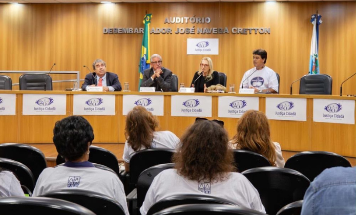 A imagem mostra um evento realizado em um auditório do Tribunal de Justiça, identificado pelas placas com o logotipo “Justiça Cidadã”. No fundo, aparece a inscrição “Auditório Desembargador José Neves Cretton”. À frente do auditório, quatro pessoas estão sentadas atrás de uma longa mesa: três homens e uma mulher. Entre eles, há uma pessoa usando uma camiseta branca com uma estampa. Sobre a mesa estão microfones, copos e outros materiais. Ao fundo, também aparecem as bandeiras do Brasil e do estado do Rio de Janeiro. Em primeiro plano, o público acompanha o encontro sentado em cadeiras do auditório. Algumas pessoas vestem camisetas brancas, e a fotografia foi registrada a partir da parte de trás da plateia, permitindo visualizar tanto os participantes da mesa quanto o ambiente da reunião.