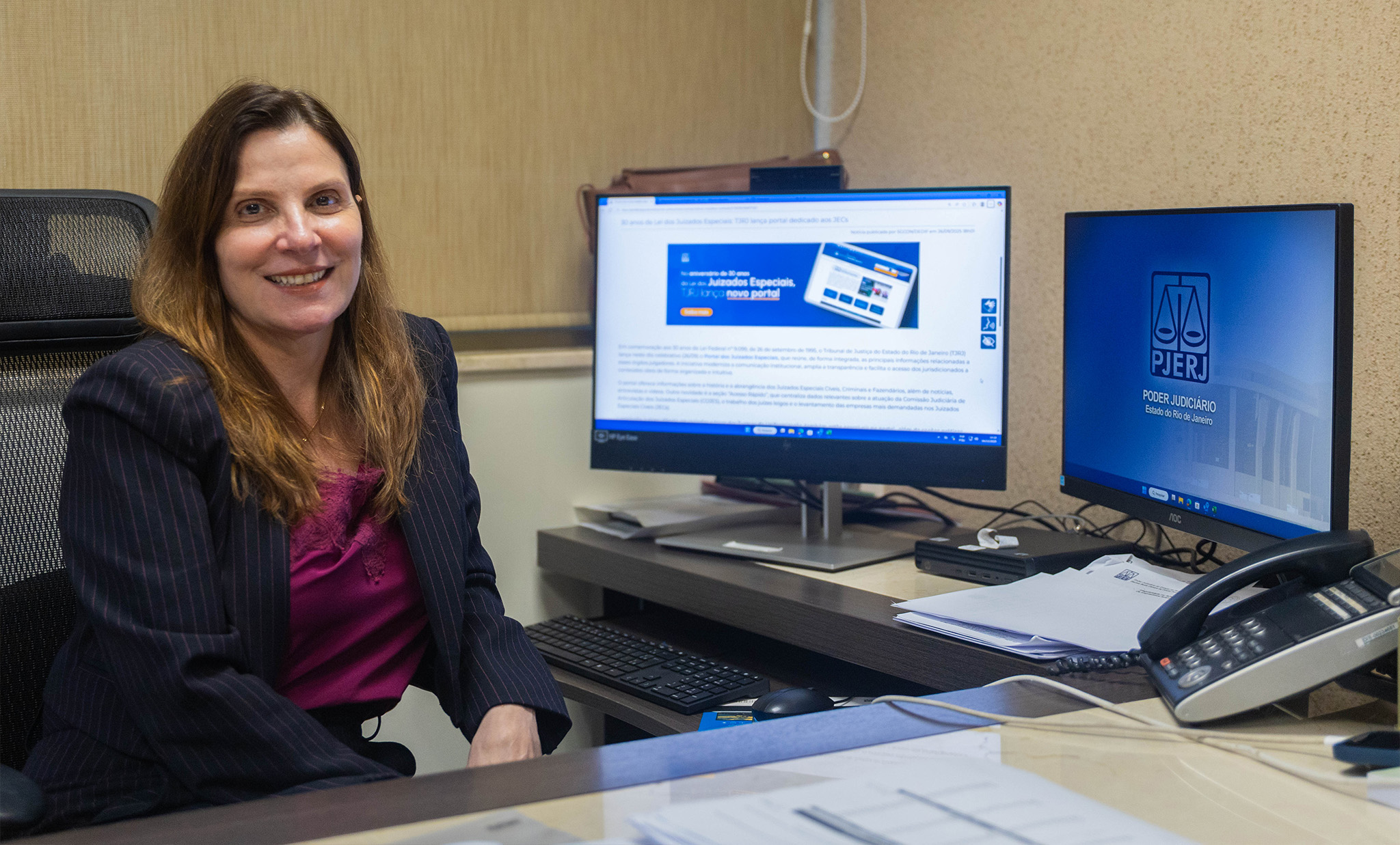 A imagem mostra a desembargadora Maria Helena Pinto Machado uma mulher sentada em um ambiente de escritório institucional, sorrindo diretamente para a câmera. Ela tem cabelos castanhos, lisos e na altura dos ombros, e veste um blazer escuro com uma blusa em tom vinho. A mesa à sua frente possui itens de trabalho, como teclado, mouse, telefone fixo e uma pilha de documentos. Ao fundo, há dois monitores de computador: um exibe uma página institucional com informações e destaque em azul, enquanto o outro apresenta o logotipo do Poder Judiciário do Estado do Rio de Janeiro (PJERJ), com o símbolo da balança.