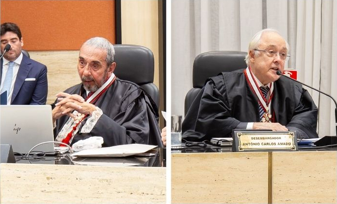 A imagem é uma montagem composta por duas fotografias lado a lado, mostrando desembargadores durante uma sessão de julgamento. À esquerda, o desembargador Mário Assis Gonçalves, de cabelos grisalhos e barba curta está sentado à bancada, usando toga e colar de mérito. Com as mãos entrelaçadas, ele observa um computador portátil aberto à sua frente. Ao fundo, outro homem de terno acompanha a sessão. À direita, o desembargador Antonio Carlos Nascimento Amado, de cabelos brancos e óculos, também veste toga e colar de mérito. Sentado à bancada, ele fala ao microfone enquanto olha para os demais participantes. À sua frente, há uma placa de identificação com o nome "Desembargador Antônio Carlos Amado", além de um copo com água e documentos sobre a mesa.