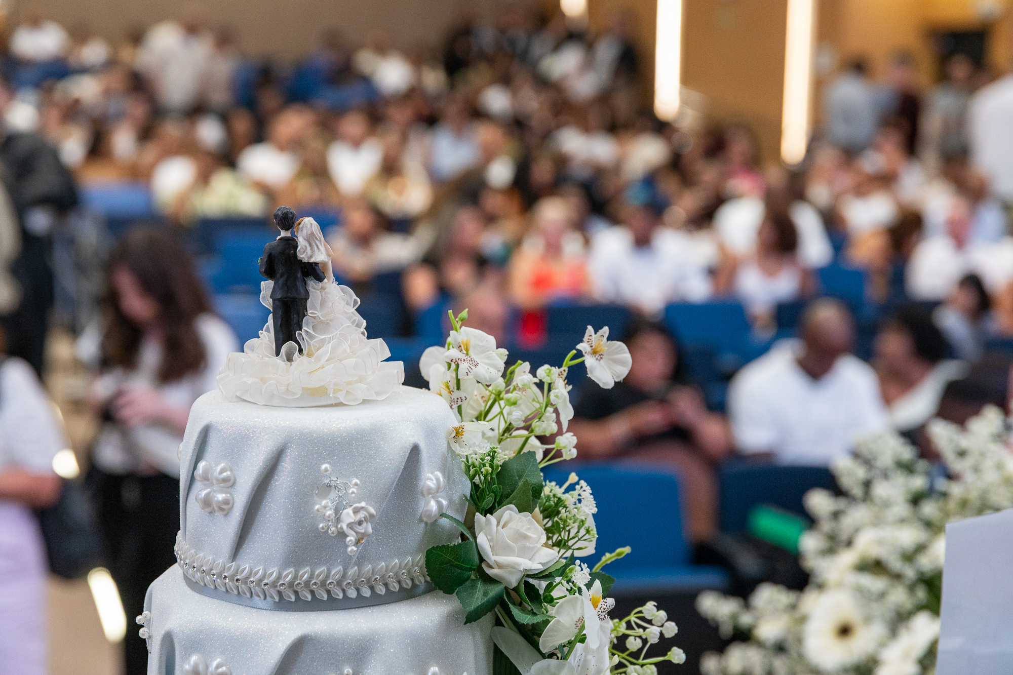 Imagem de um bolo de casamento branco, com três andares e no topo um casal de noivos. No fundo da foto, a plateia do auditório formada por casais