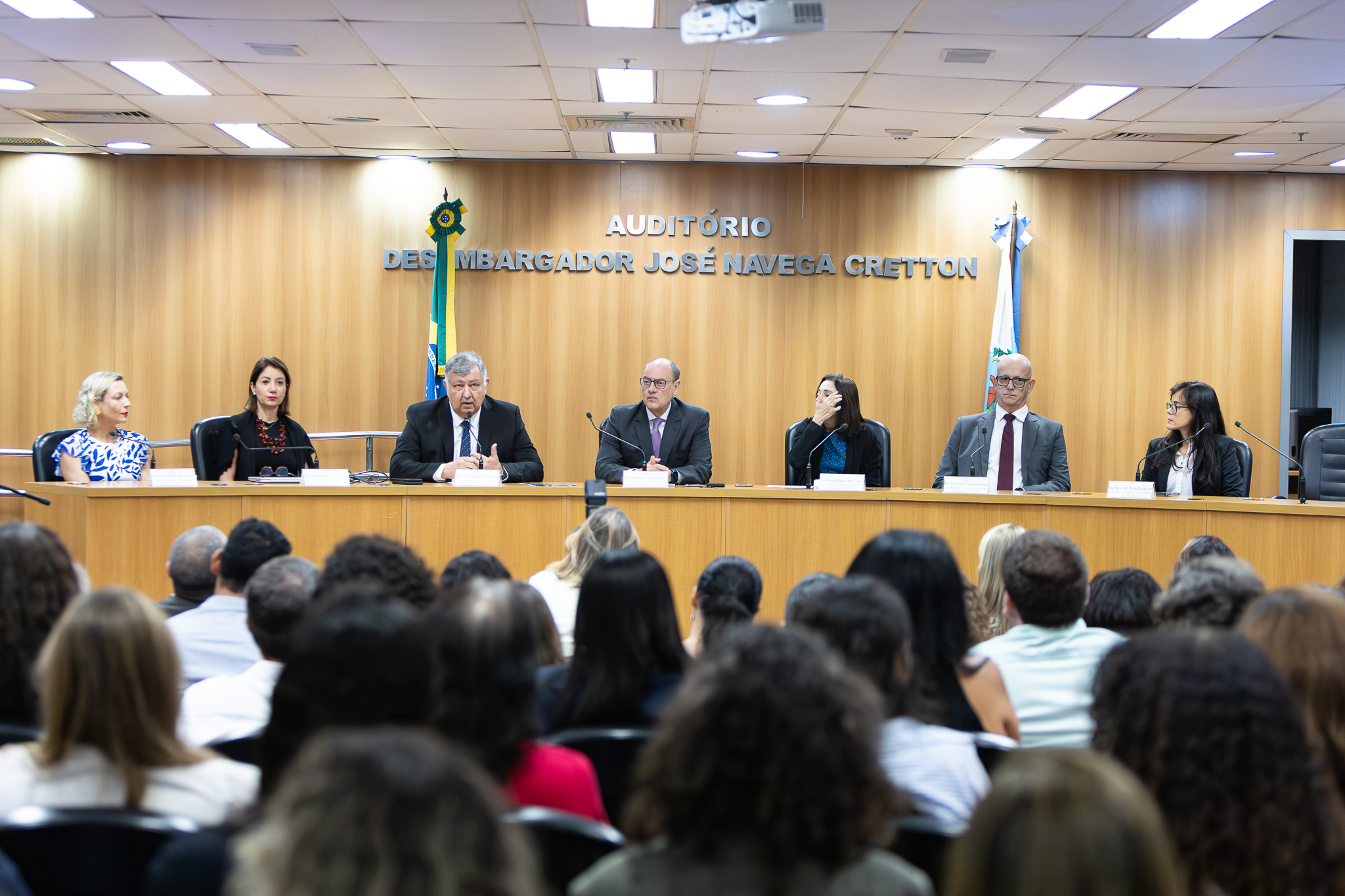 Os participantes estão sentados lado a lado atrás de uma longa mesa de madeira, cada um com microfone e identificação, sugerindo tratar-se de uma mesa de debate. No centro, um homem de terno escuro fala ao microfone, com expressão séria. Os demais integrantes — homens e mulheres vestidos formalmente — demonstram atenção, alguns olhando para o orador, outros para o público. Ao fundo, na parede de madeira, está escrito “Auditório Desembargador José Navega Cretton”, identificando o local. Também são visíveis bandeiras oficiais. Em primeiro plano, aparece a plateia, com diversas pessoas sentadas e voltadas para a mesa, acompanhando a apresentação. 