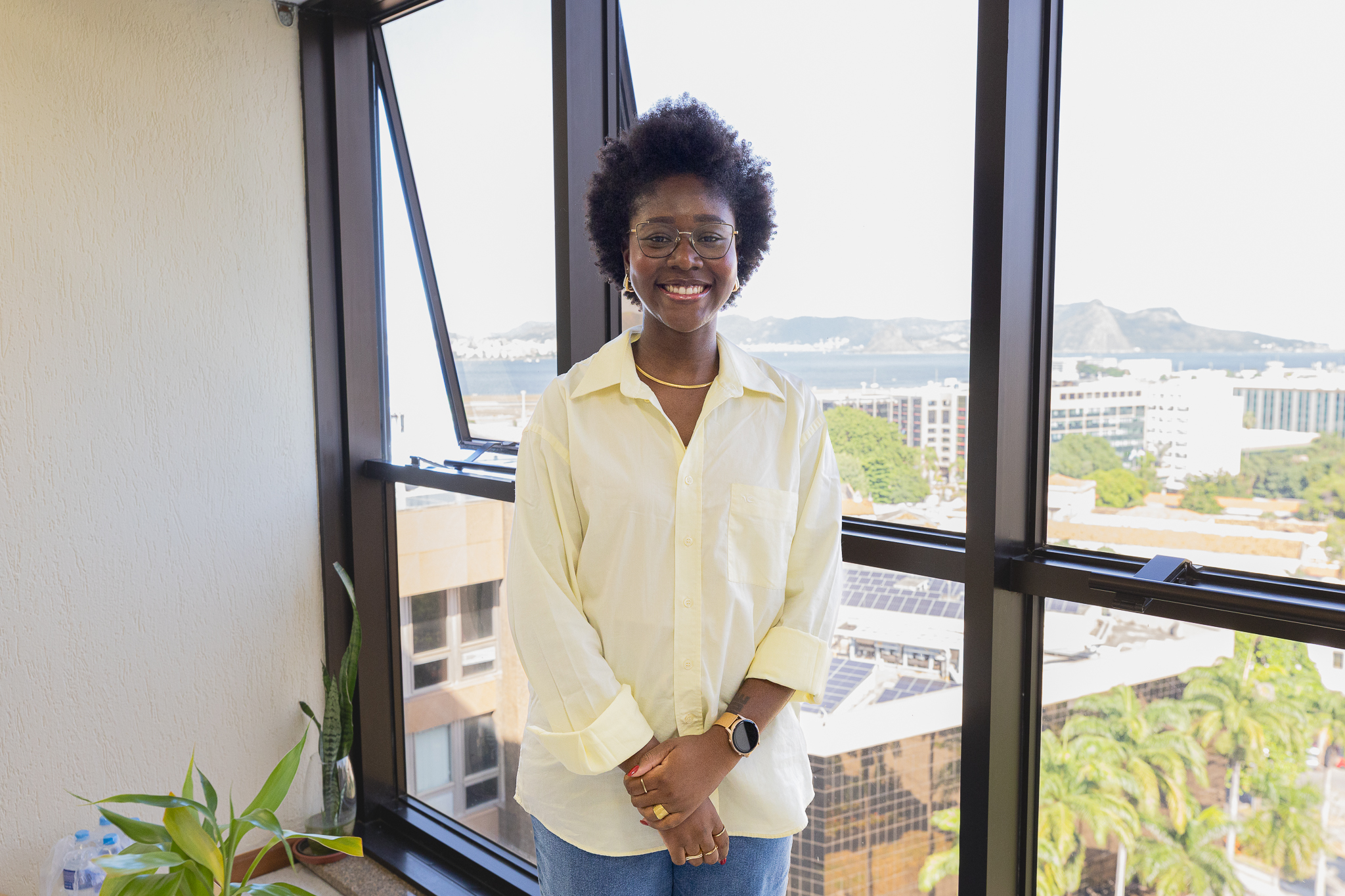 A imagem mostra uma mulher posando para a foto em pé com uma janela atrás.