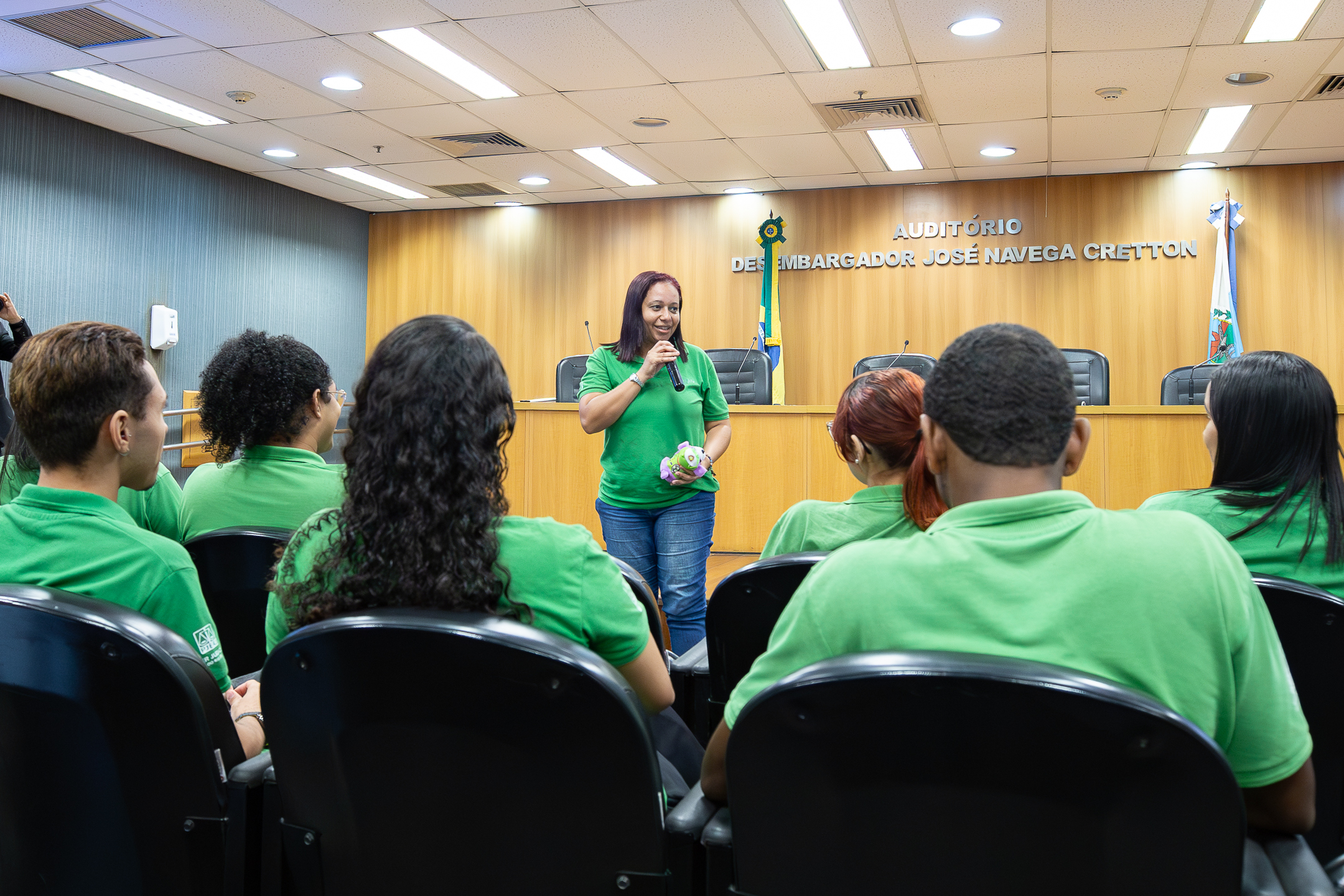 A imagem mostra uma atividade em um auditório institucional. Em primeiro plano, participantes sentados em cadeiras assistem atentamente a uma mulher que está à frente, conduzindo a fala. Ela está vestida com camisa verde, segura um microfone. Os demais participantes, também com camisas verdes, estão voltados para ela. O ambiente é formal, com mesa de autoridades ao fundo e bandeiras posicionadas atrás. Na parede, lê-se “Auditório Desembargador José Navega Cretton”.