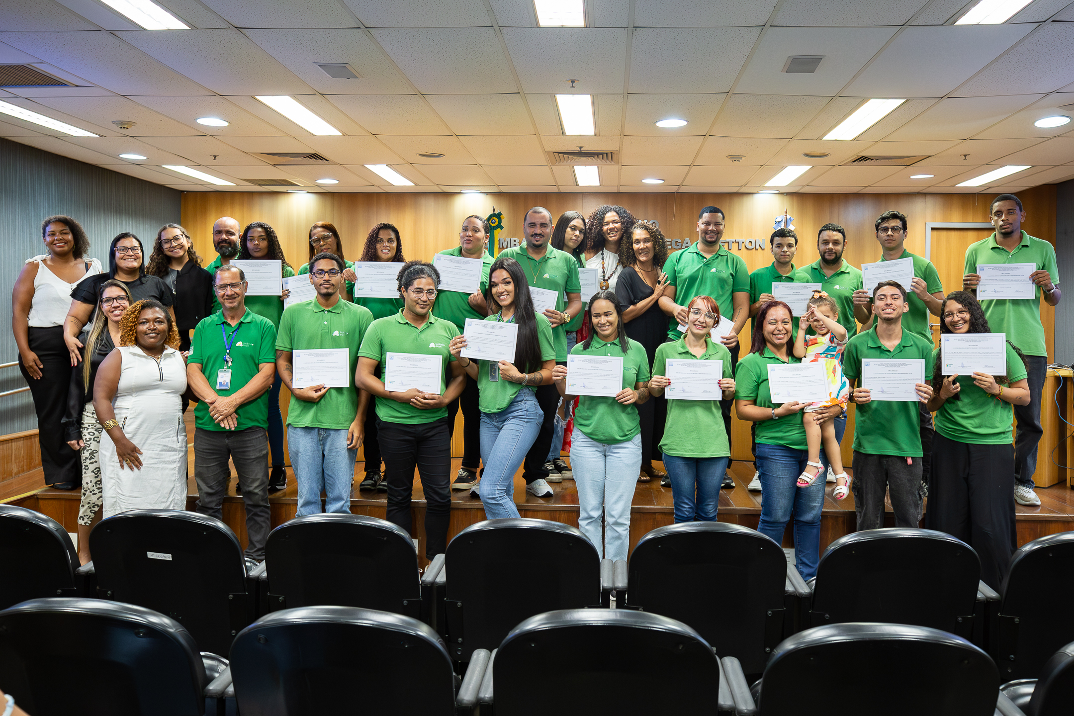 A imagem mostra um grupo numeroso de pessoas posando para uma foto em um auditório. Elas estão organizadas em duas fileiras, sobre um pequeno palco, e a maioria veste camisas verdes. Muitos dos participantes seguram certificados, exibindo-os para a câmera com expressão de satisfação. Há diversidade entre os integrantes do grupo, com homens e mulheres de diferentes idades. À esquerda, algumas pessoas vestem roupas diferentes das camisas verdes. O ambiente é um auditório institucional, com cadeiras em primeiro plano e parede ao fundo em madeira, onde se vê parcialmente um símbolo ou identificação do local. 