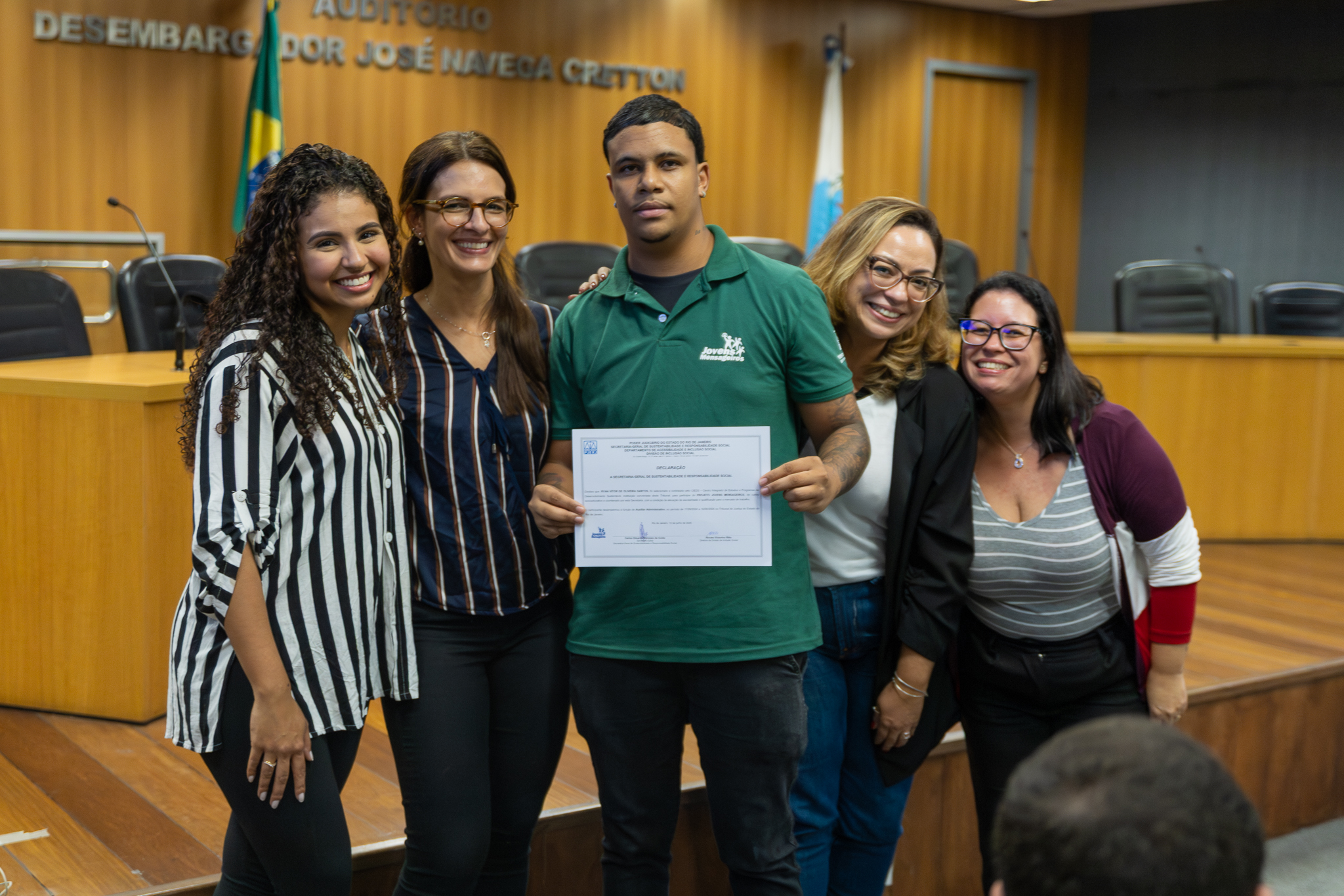 A imagem mostra cinco pessoas posando para uma fotografia em um auditório durante uma cerimônia. No centro da cena, um jovem veste uma camisa polo verde e segura um certificado, símbolo da conclusão do curso. Ao seu redor, quatro mulheres sorriem para a câmera, posicionadas de forma próxima e acolhedora, demonstrando apoio e celebração pela conquista. Duas delas usam blusas listradas, uma veste blazer preto sobre blusa clara e outra usa cardigan vinho sobre camiseta listrada. Ao fundo, é possível observar a mesa principal do auditório, cadeiras institucionais, bandeiras e o revestimento de madeira da parede. Na parte superior da imagem aparece a identificação do auditório. 