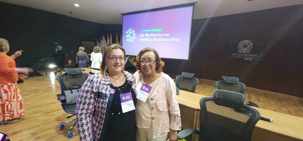 Duas mulheres posam juntas tendo ao fundo uma mesa grande, um telão e uma pessoa de costa. A imagem sugere uma foto de integração pós seminário. Estão na foto a juíza Claudia Marcia Vidal, do TJRJ, e a desembargadora Joanice Guimarães, do TJBA