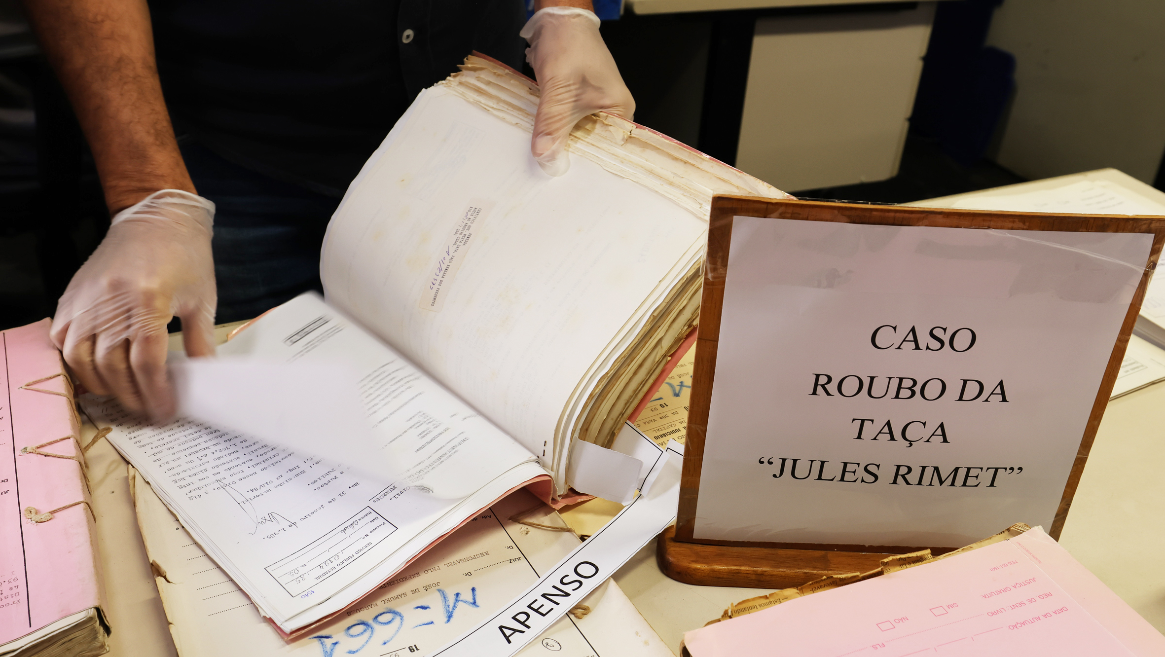 A imagem mostra um conjunto de processos judiciais antigos sobre uma mesa, com um profissional usando luvas descartáveis para manusear cuidadosamente um dos volumes. À direita, há uma placa identificando o material como "Caso Roubo da Taça 'Jules Rimet'". Os documentos apresentam folhas amareladas pelo tempo, evidenciando seu valor histórico e de preservação. Ao redor, outros processos e pastas de diferentes cores estão empilhados, compondo um cenário típico de consulta a acervos arquivísticos. A cena destaca o trabalho de conservação e pesquisa de documentos relacionados a um dos casos mais emblemáticos da história do futebol brasileiro.