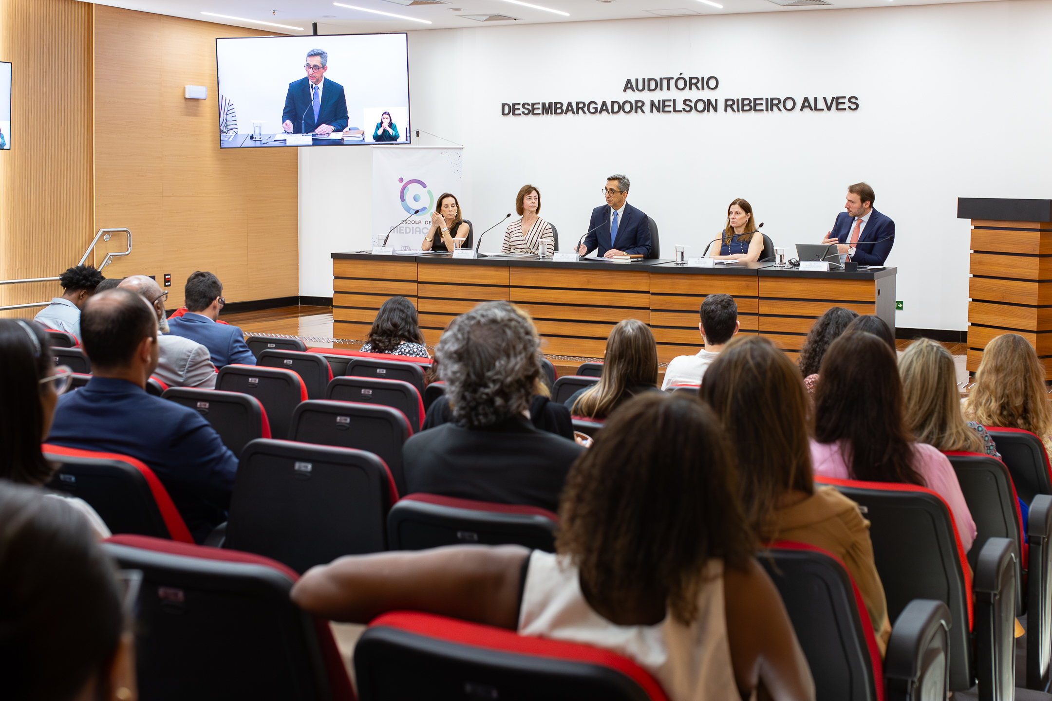 A imagem mostra um auditório durante a realização de um evento institucional. Em primeiro plano, o público está sentado em cadeiras estofadas, voltado para a mesa principal. À frente, cinco pessoas compõem uma mesa de debate, cada uma com microfone e identificação. Um dos participantes, ao centro, fala enquanto os demais escutam atentamente. O ambiente é moderno, com paredes claras e mobiliário em madeira. Na parede ao fundo, está escrito “Auditório Desembargador Nelson Ribeiro Alves”, indicando o local do evento. À esquerda, uma tela exibe a transmissão de um participante remoto, acompanhado de uma intérprete de Libras.   