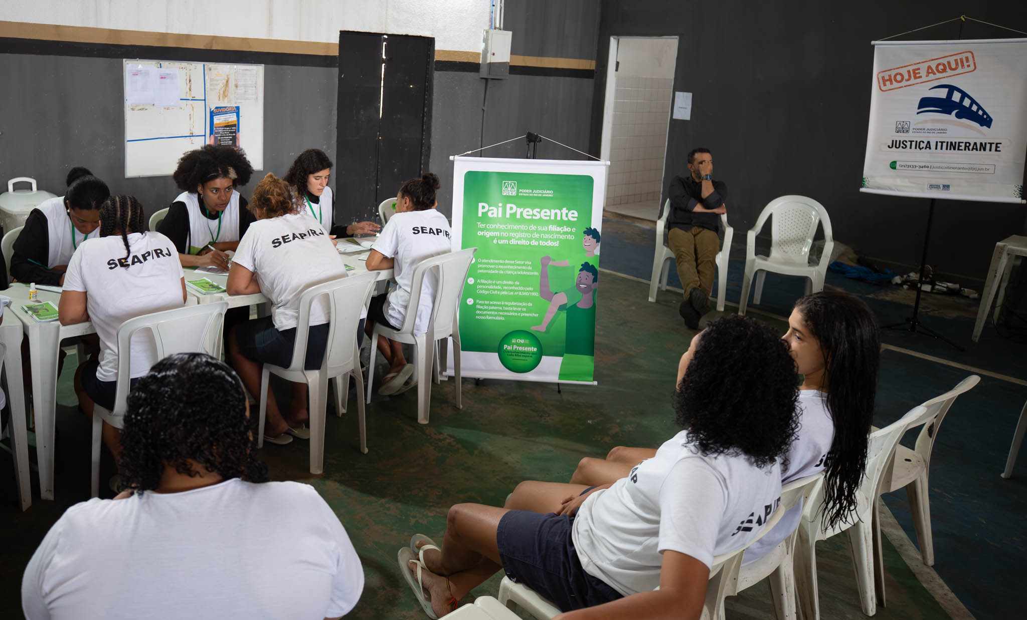 A imagem mostra detentas sendo atendidas em mesas no pátio com banners da Justiça Itinerante e do Pai Presente no fundo