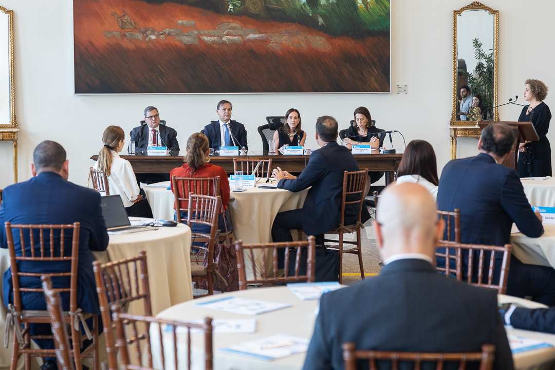 Em um amplo salão com mesas redondas espalhadas, há uma mesa retangular central com quatro pessoas, dois homens e duas mulheres, conduzindo uma reunião. 