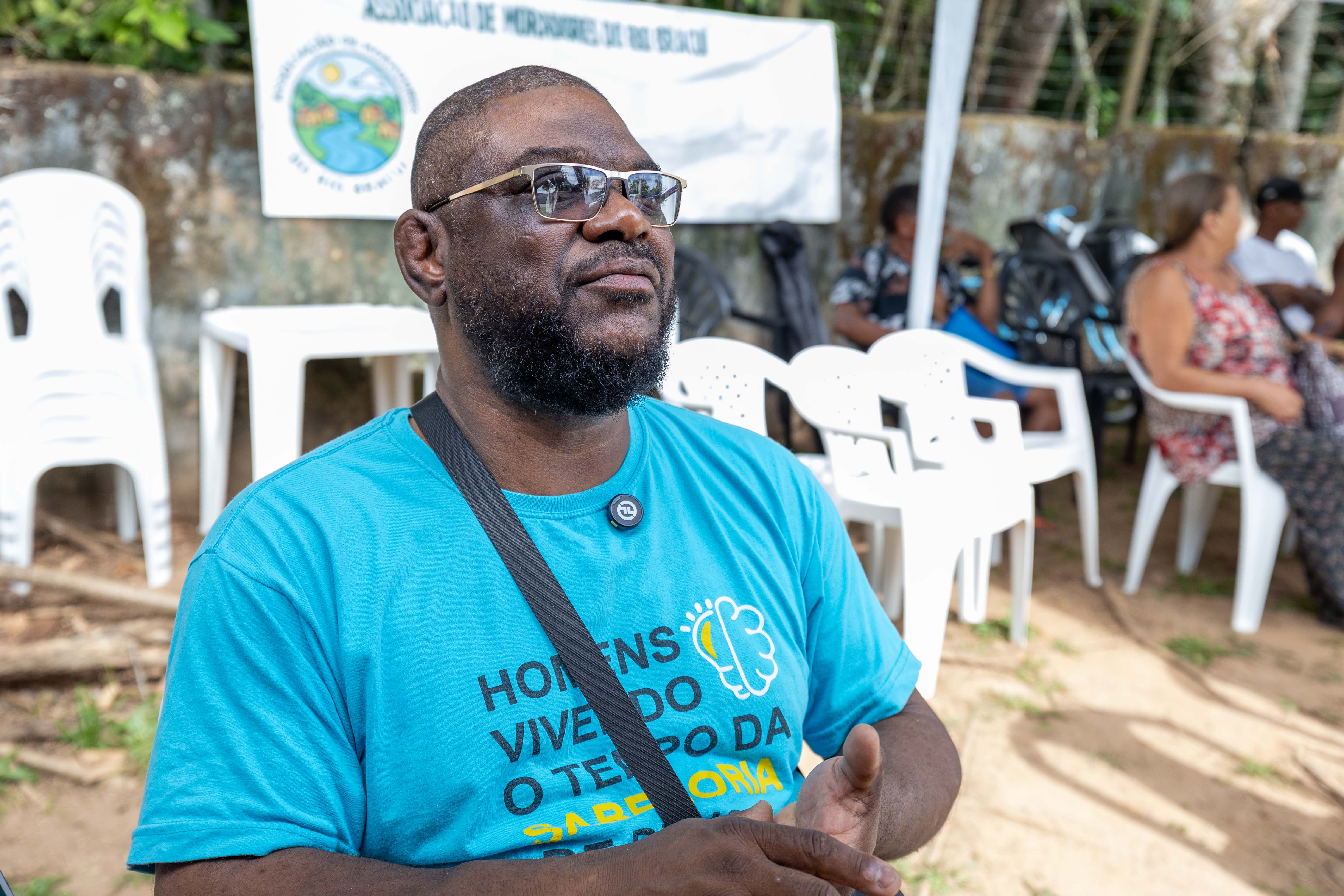 Em uma área externa com cadeiras plásticas brancas e tendas ao fundo, um homem negro usa óculos e camiseta azul com uma mensagem estampada sobre memória e direitos. Ele aparece em primeiro plano, olhando para cima com expressão atenta e as mãos unidas à frente do corpo. Uma faixa da associação de moradores está posicionada ao fundo, enquanto outras pessoas permanecem sentadas sob a tenda em um espaço comunitário arborizado.