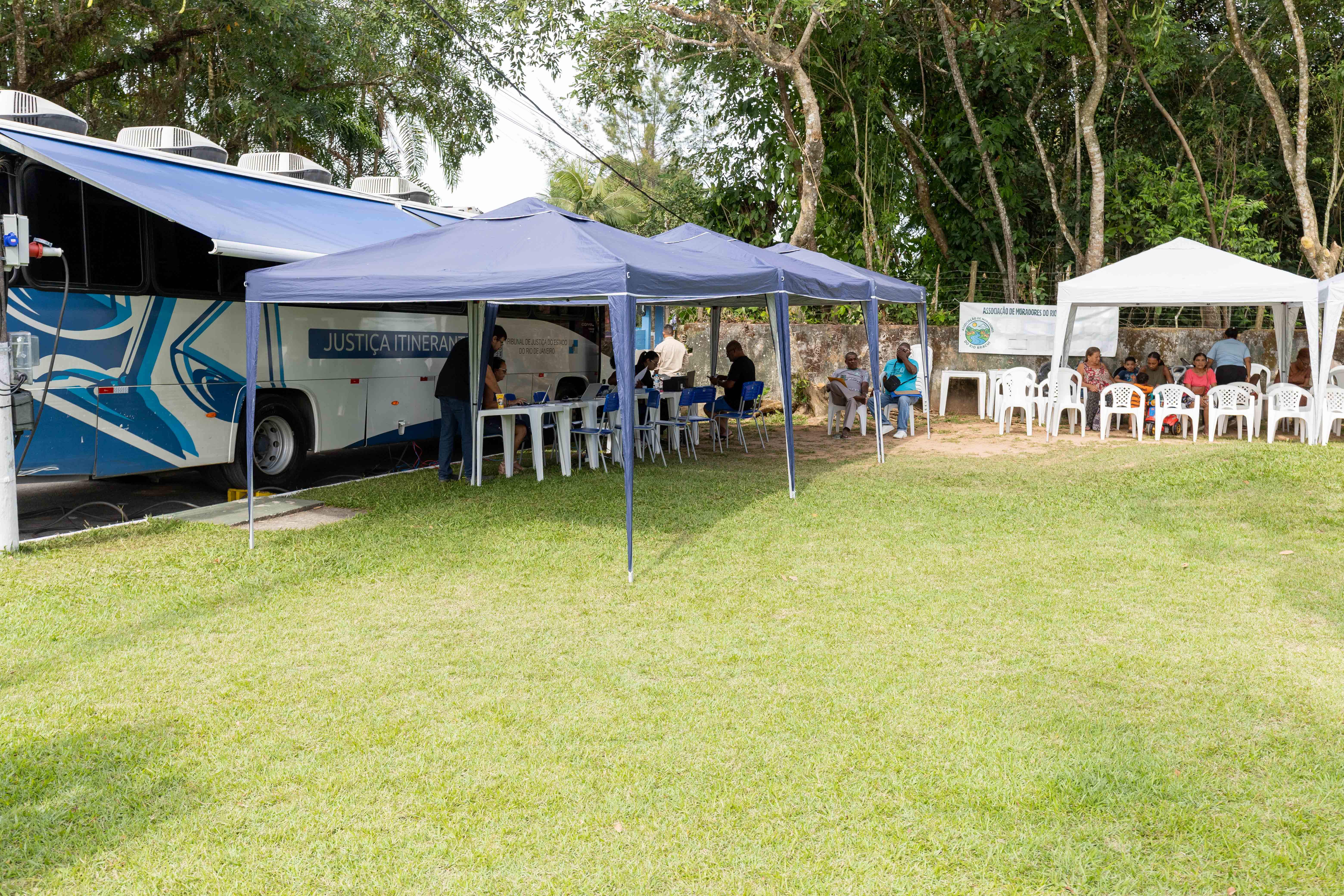 Em uma área gramada e arborizada, tendas azuis e brancas foram montadas para atendimento ao público ao lado de um ônibus da Justiça Itinerante. Sob as tendas azuis, mesas e cadeiras acomodam atendentes e moradores durante os serviços prestados. À direita, pessoas aguardam sentadas em cadeiras plásticas brancas sob uma tenda branca. Ao fundo, árvores altas cercam o espaço, criando um ambiente aberto e sombreado. Uma placa de associação de moradores também aparece próxima à área de atendimento.