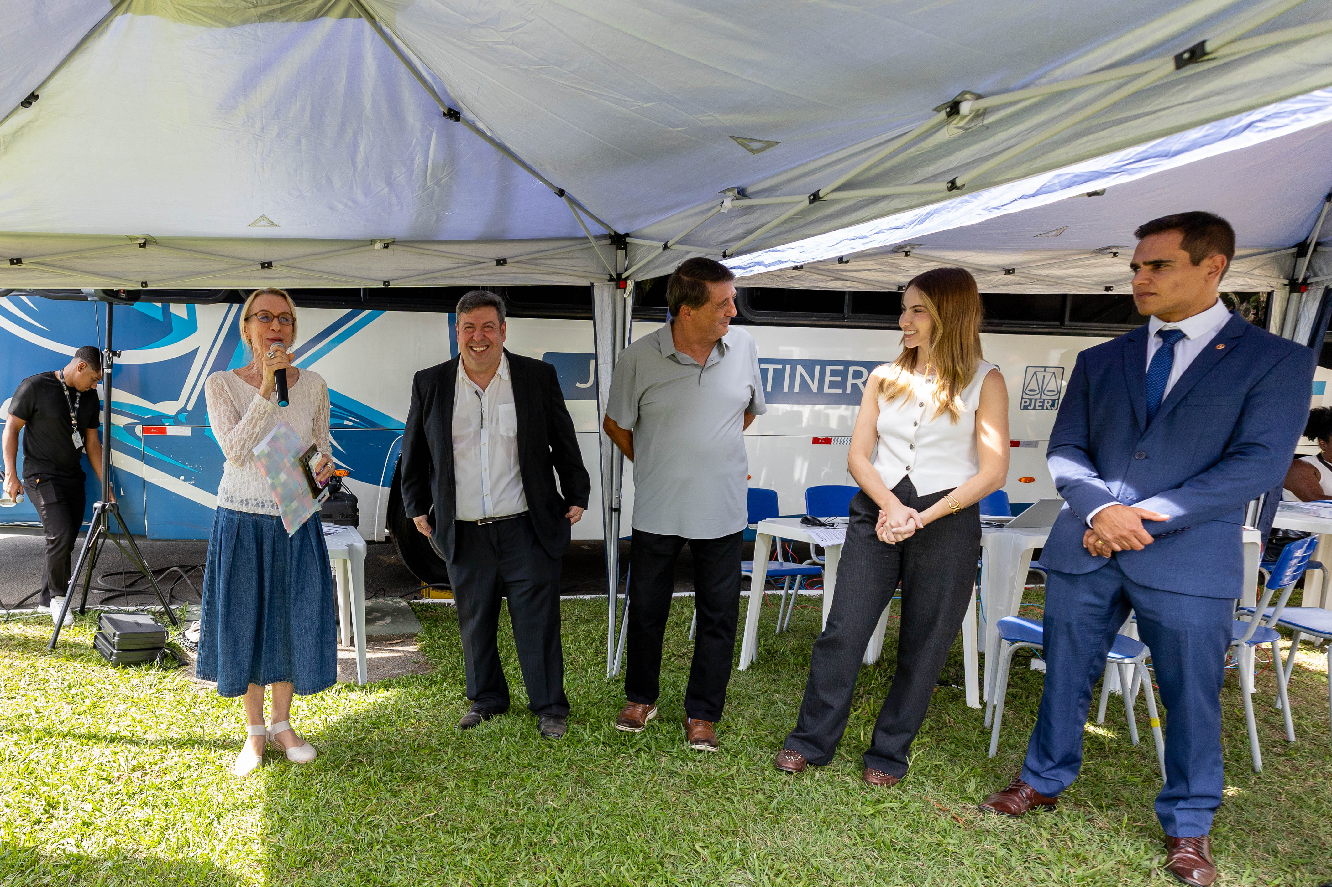 Em uma área gramada, sob tendas brancas montadas ao ar livre, cinco pessoas participam de um evento institucional diante de um ônibus da Justiça Itinerante do Poder Judiciário do Estado do Rio de Janeiro (PJERJ). À esquerda, a desembargadora Cristina Tereza Gaulia, uma mulher de cabelos claros e óculos fala ao microfone enquanto segura uma pasta colorida. Ao lado dela, dois homens observam sorrindo, um deles vestindo terno escuro e outro usando camisa polo cinza. Mais à direita, uma mulher de blusa branca sem mangas e calça preta sorri enquanto conversa com o grupo. Na extremidade direita, um homem de terno azul permanece em posição formal, com as mãos à frente do corpo. Ao fundo, há mesas, cadeiras e equipamentos de som sob as tendas, além de uma pessoa trabalhando próxima aos aparelhos de áudio.