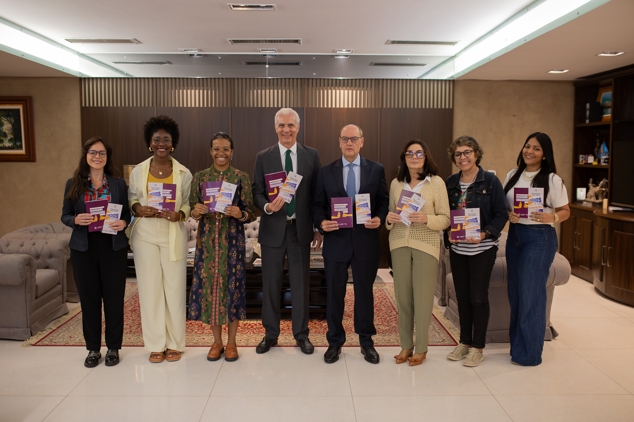 A imagem mostra oito pessoas posando lado a lado em um ambiente interno de aspecto sofisticado e institucional. Todas estão em pé, sorrindo para a câmera e segurando exemplares de uma cartilha de capa roxa. No centro do grupo, dois homens vestem ternos escuros e gravatas. Ao redor deles, estão seis mulheres com roupas em estilos variados, incluindo blazer, vestido estampado, conjunto claro, jeans e tricô. Algumas usam óculos. O ambiente possui decoração elegante, com sofás cinza, tapete estampado, mesa de centro e painéis de madeira ao fundo. À direita, há estantes com objetos decorativos e livros. A iluminação é clara e uniforme, reforçando o caráter formal do encontro ou lançamento institucional.
