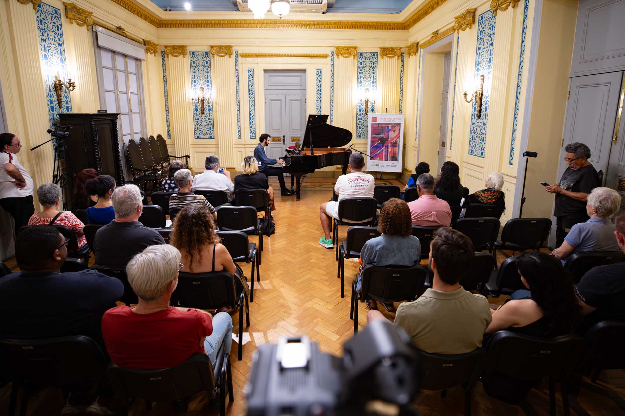 Fotografia mostra uma apresentação musical realizada em um salão histórico e ornamentado. Ao centro, um pianista está sentado diante de um piano de cauda preto, tocando para o público. À frente dele, há um microfone direcionado ao instrumento. Cerca de 30 pessoas estão sentadas em cadeiras dispostas em fileiras, acompanhando a apresentação. Os espectadores aparecem de costas e de perfil, alguns atentos ao músico e outros registrando o momento com celulares. O salão possui decoração clássica, com paredes em tons claros, detalhes dourados, colunas e painéis ornamentais em azul e branco. Arandelas iluminam as paredes, enquanto o piso de madeira apresenta um desenho em espinha de peixe. À direita do piano, há um painel vertical com informações sobre o evento.