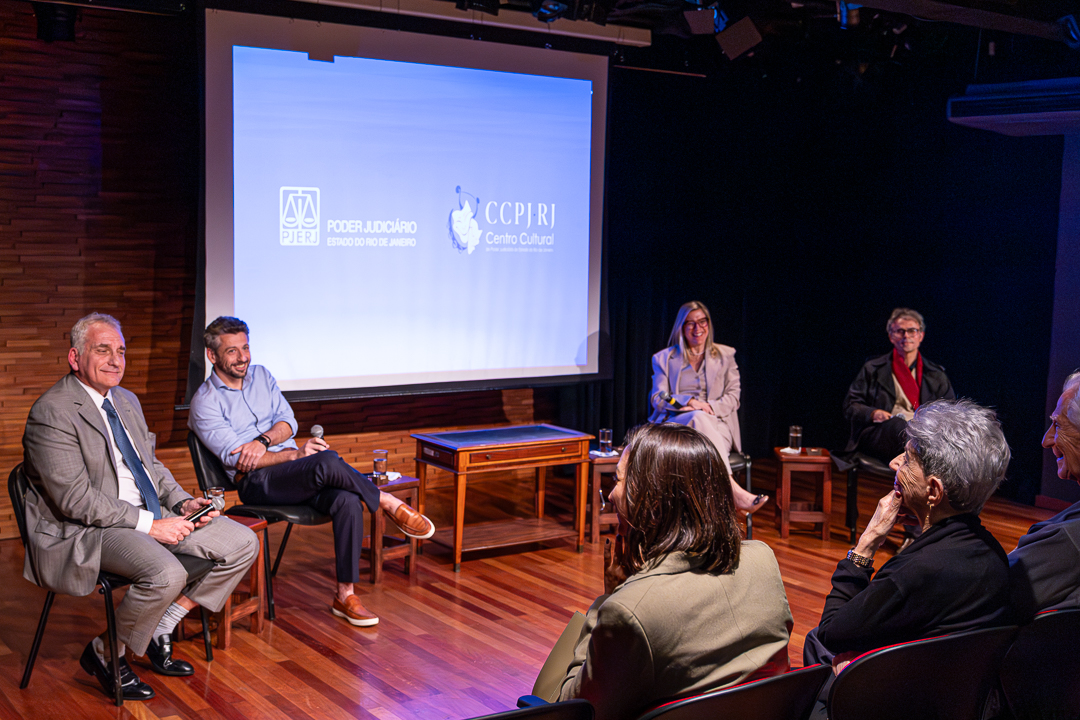 Participantes do evento, sentado em cadeiras em semicírculo no palco da sala, com telão ao fundo e de frente para a plateia