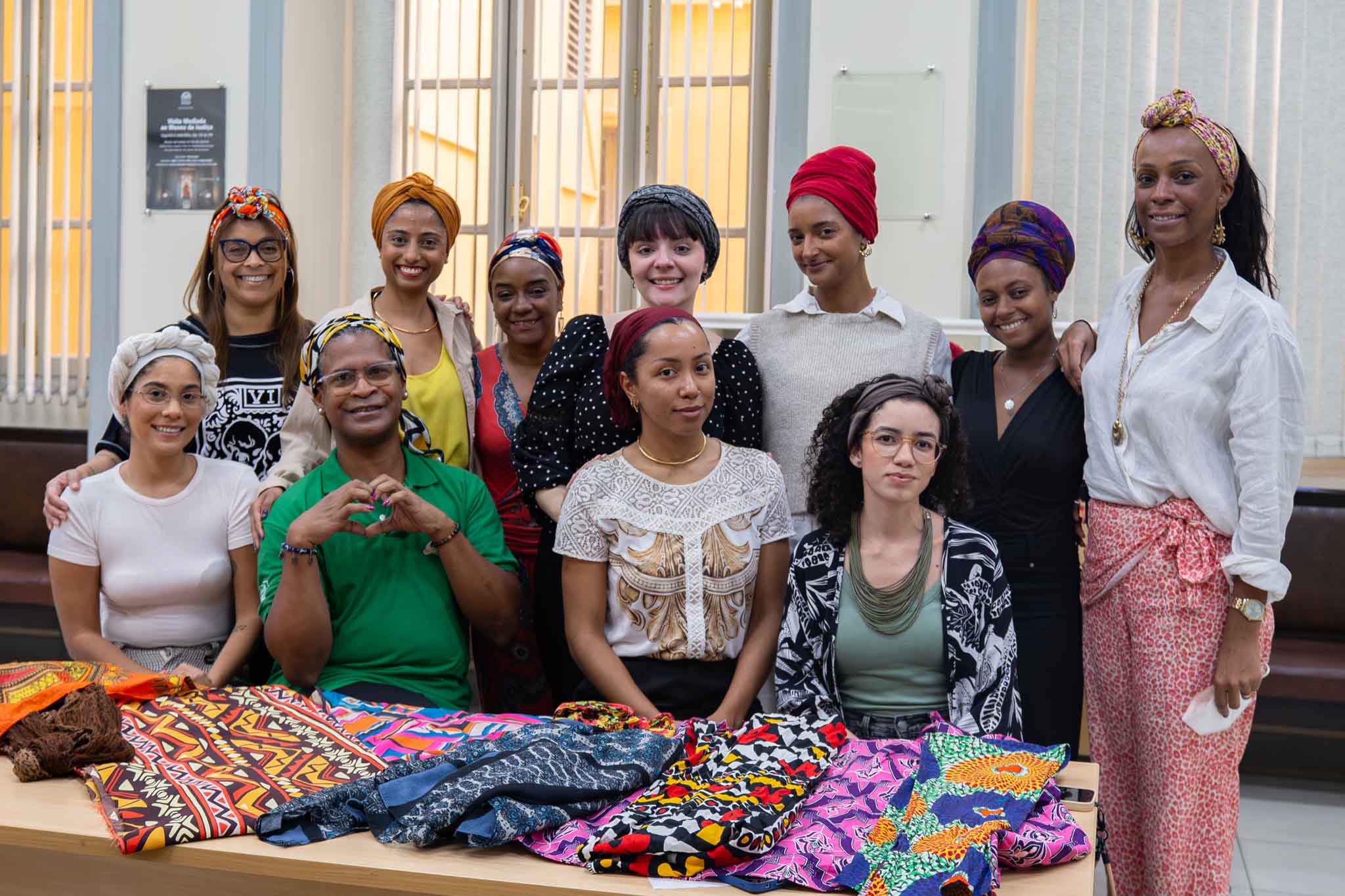 A imagem mostra um grupo de 11 pessoas reunidas em um ambiente interno, posando para uma fotografia atrás de uma mesa coberta por tecidos estampados e turbantes coloridos. A maioria dos participantes sorri para a câmera e utiliza turbantes ou lenços amarrados na cabeça em diferentes cores e estilos, como vermelho, amarelo, laranja, roxo e estampas multicoloridas. Na primeira fila, quatro pessoas estão sentadas e uma está em pé, com as mãos formando um coração. Na segunda fila, outras sete pessoas permanecem em pé, lado a lado, algumas com os braços apoiados nos ombros dos colegas, transmitindo um clima de integração e acolhimento. Sobre a mesa, estão expostos diversos tecidos com estampas vibrantes e de inspiração africana, em tons de laranja, rosa, azul, preto, amarelo e verde, utilizados na oficina.