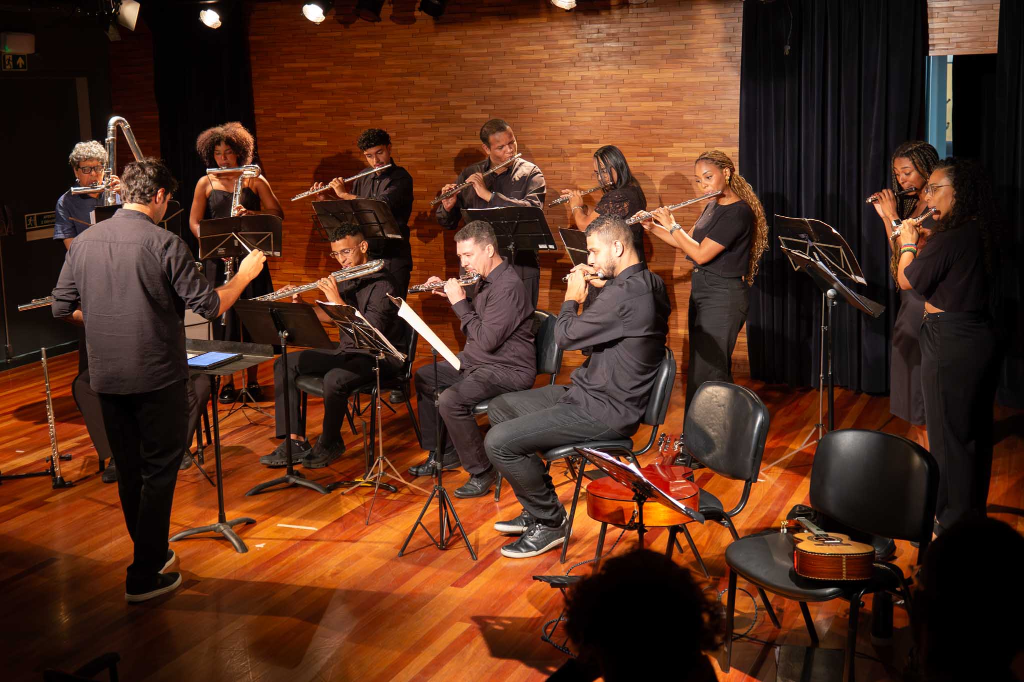 A imagem mostra uma apresentação musical em um palco de ambiente fechado, com piso e paredes revestidos em madeira, criando uma atmosfera acolhedora e elegante. No centro da cena, um maestro, de costas para a câmera, conduz um grupo de músicos durante a execução da peça. A orquestra é formada por homens e mulheres vestidos predominantemente de preto, distribuídos entre músicos sentados e em pé. A maioria toca flautas de diferentes tamanhos, enquanto partituras estão apoiadas em estantes à frente dos instrumentistas. A iluminação do palco destaca os músicos e os instrumentos, valorizando o momento da performance.