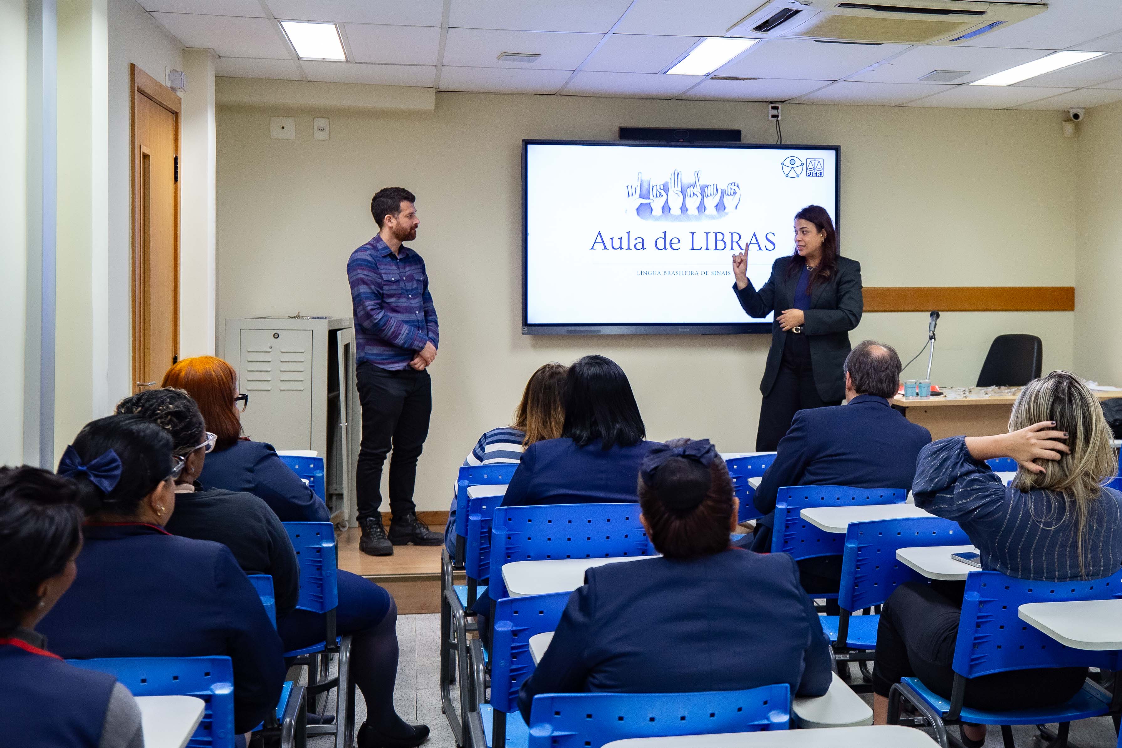Foto mostra um homem e uma mulher à frente de uma turma de alunos sentados em cadeiras azuis. Ao fundo, um telão destaca a frase Aula de Libras