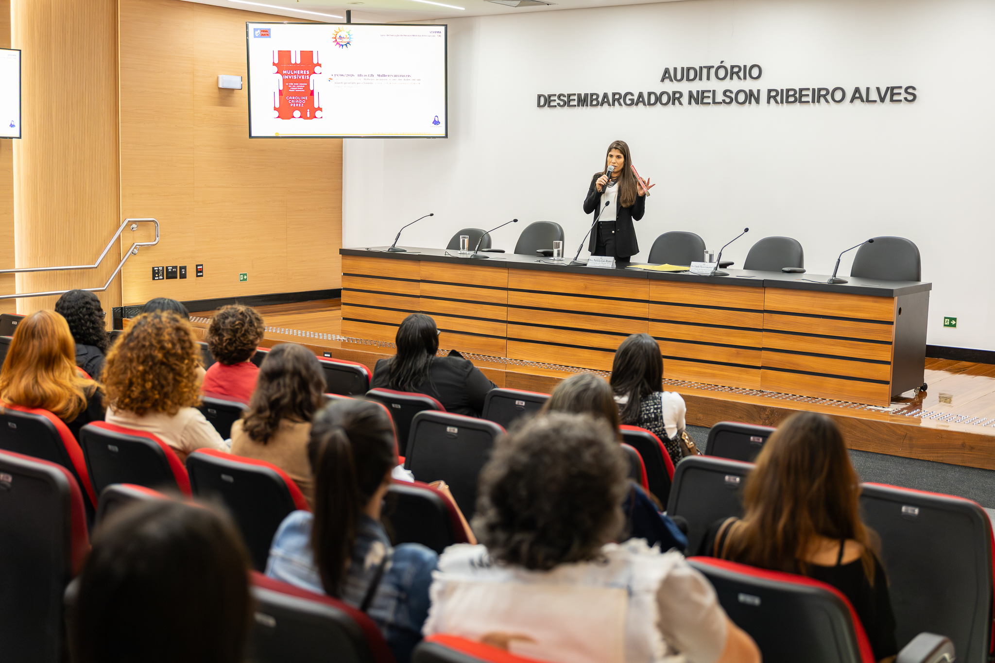 A imagem mostra uma palestra realizada em um auditório. Em primeiro plano, o público ocupa fileiras de cadeiras estofadas nas cores preta e vermelha, voltadas para o palco. Os participantes acompanham atentamente a apresentação. No centro do auditório, uma mulher está em pé, segurando um microfone enquanto fala para a plateia. Ela gesticula com uma das mãos, demonstrando interação e dinamismo durante a exposição. Atrás dela, há uma mesa longa com assentos e microfones, preparada para debates ou composições de mesa-redonda. Na parede ao fundo, destaca-se a inscrição “Auditório Desembargador Nelson Ribeiro Alves”. Acima do palco, uma tela exibe a capa do livro “Mulheres Invisíveis”, acompanhada de informações sobre o evento, indicando que a atividade está relacionada a uma palestra, lançamento ou debate sobre a obra.