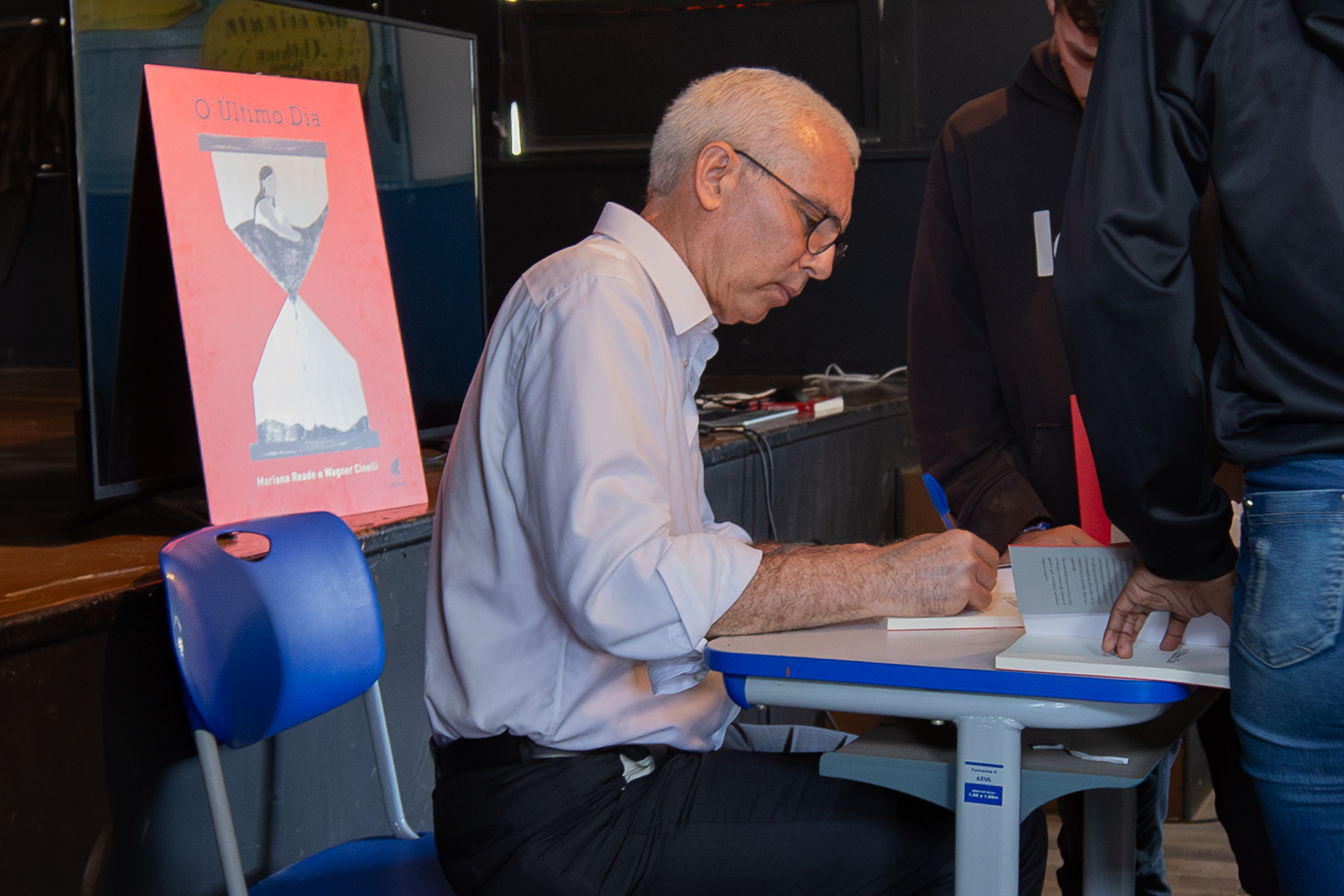 A imagem mostra o desembargador Wagner Cinelli sentado em uma cadeira escolar azul, assinando um livro apoiado sobre uma pequena mesa. Ele veste camisa clara de mangas compridas e usa óculos, concentrado na dedicatória que escreve. Ao lado dele, há outras pessoas em pé, uma delas segurando um exemplar aberto, aguardando. À esquerda da cena, há um cartaz em tom rosado com a capa de um livro intitulado “O Último Dia”, ilustrado com uma ampulheta.