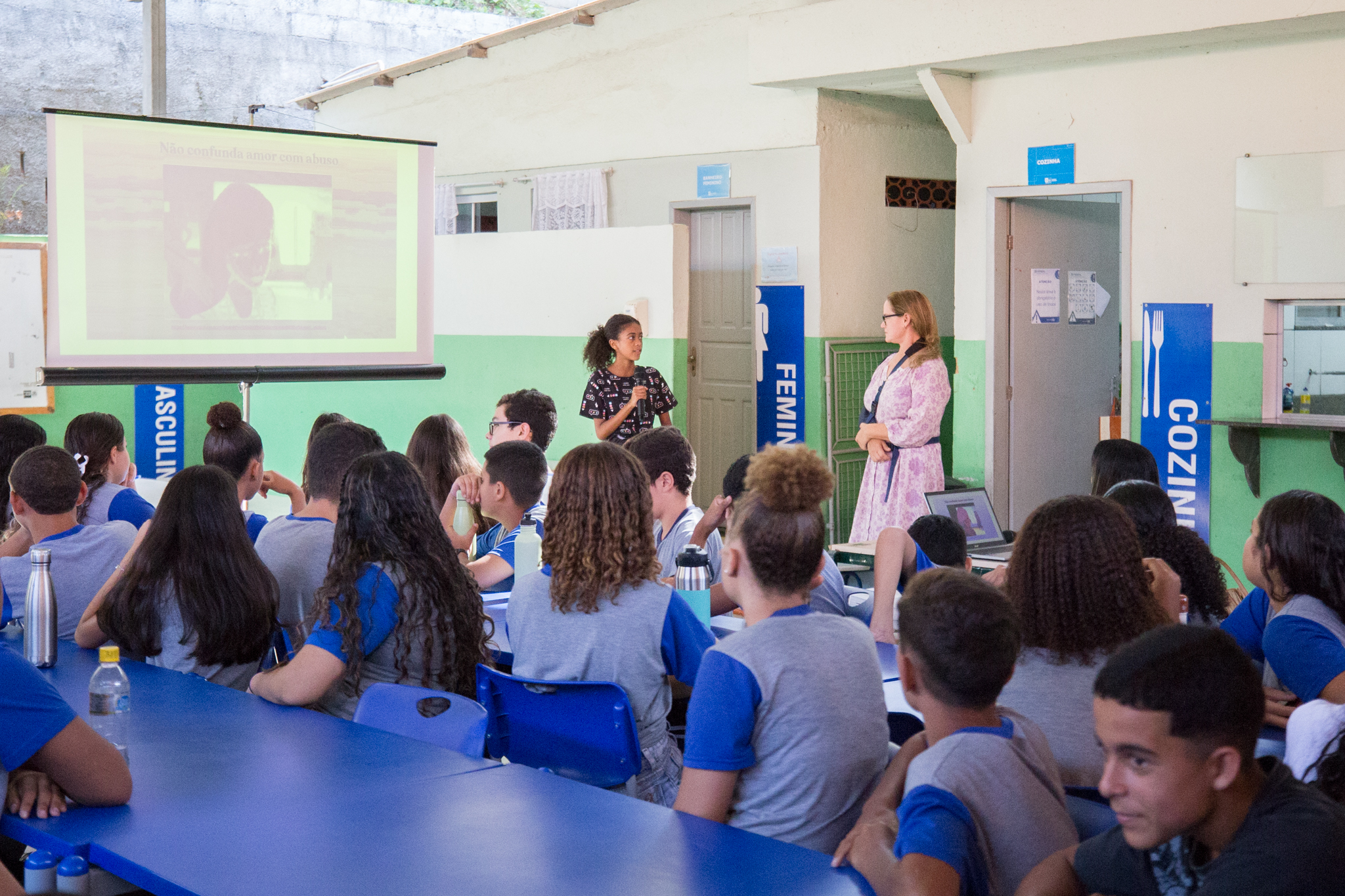 A imagem mostra uma atividade educativa com estudantes em um espaço semelhante a um refeitório escolar. Os alunos, usando uniformes em tons de azul e cinza, estão sentados em mesas compridas, voltados para a frente e atentos à apresentação. No centro, uma adolescente está em pé, segurando um microfone e falando para os colegas, assumindo o papel de participante ativa da atividade. Ao lado, juíza Katerine Jatahy — vestindo um vestido claro com estampa e cinto — observa a fala da estudante com atenção, em postura de mediação. À esquerda, um telão projeta um slide com a frase “Não confunda amor com abuso”, acompanhado da imagem de uma pessoa, indicando que o tema da atividade está relacionado à conscientização sobre relacionamentos abusivos e violência.