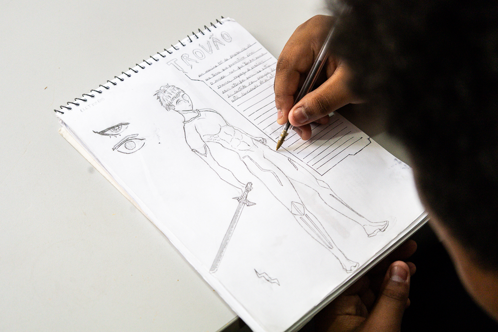   Foto mostra mão de jovem segurando caneta e desenhando em bloco de fundo branco a imagem de um super-herói