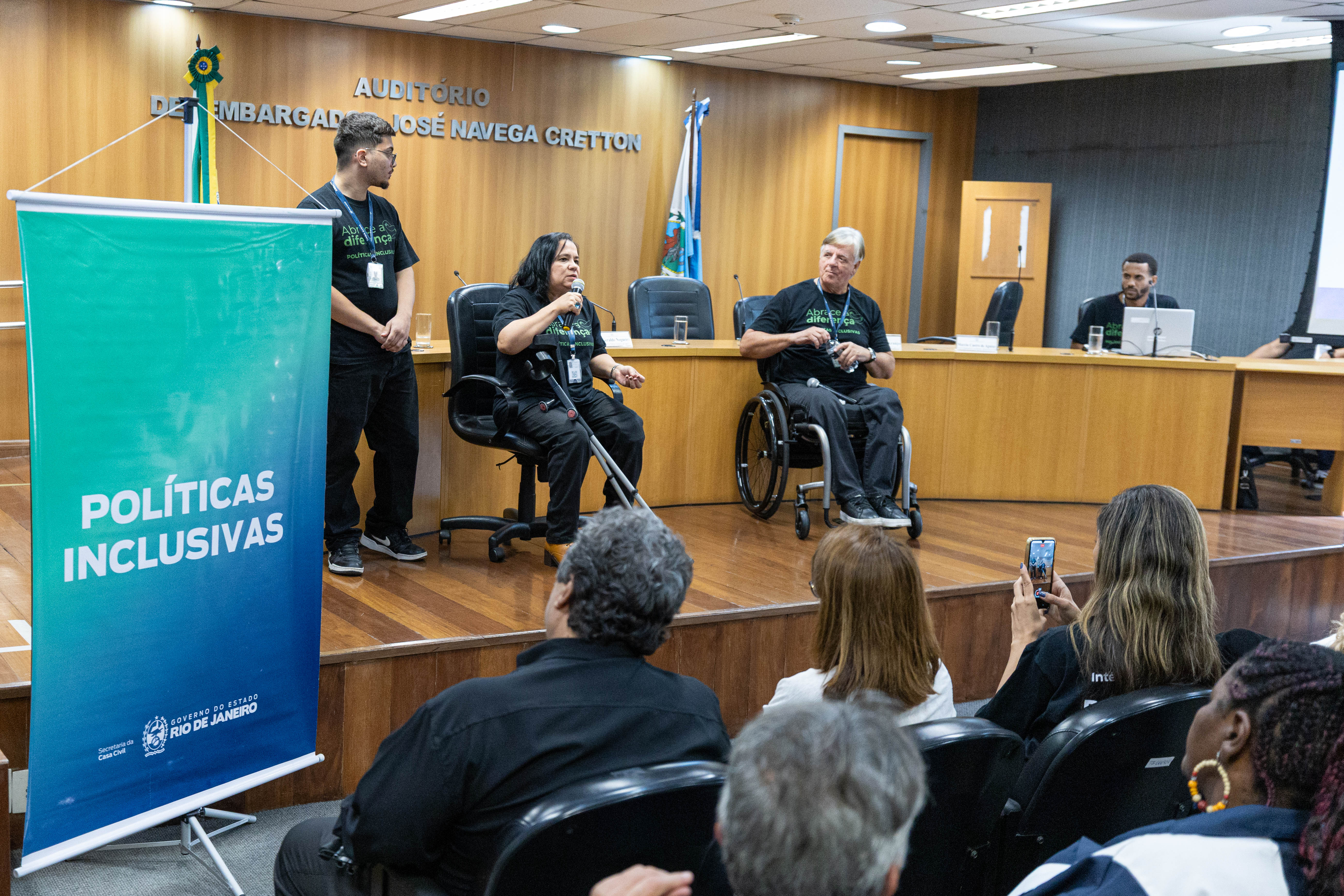 Imagem do auditório Desembargador José Navega Cretton. Ao fundo os convidados do evento falando para a plateia 