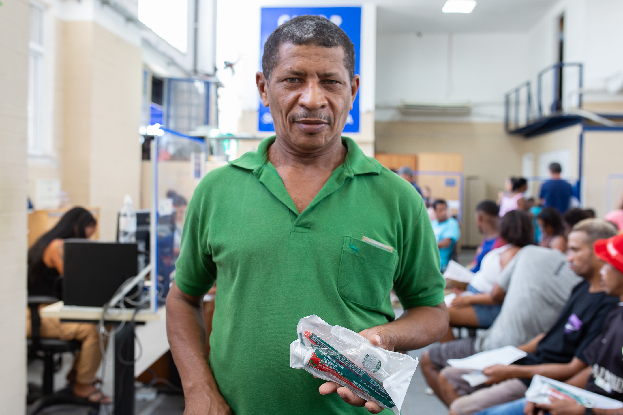 A imagem mostra um homem em primeiro plano, em pé, segurando um pequeno kit embalado, contendo itens de saúde ou higiene. Ele veste uma camisa verde e olha diretamente para a câmera, com expressão séria. Ao fundo, o ambiente parece ser uma unidade de atendimento, com várias pessoas sentadas aguardando, algumas segurando papéis ou documentos. Há mesas com computadores e divisórias, indicando atendimento organizado ao público.