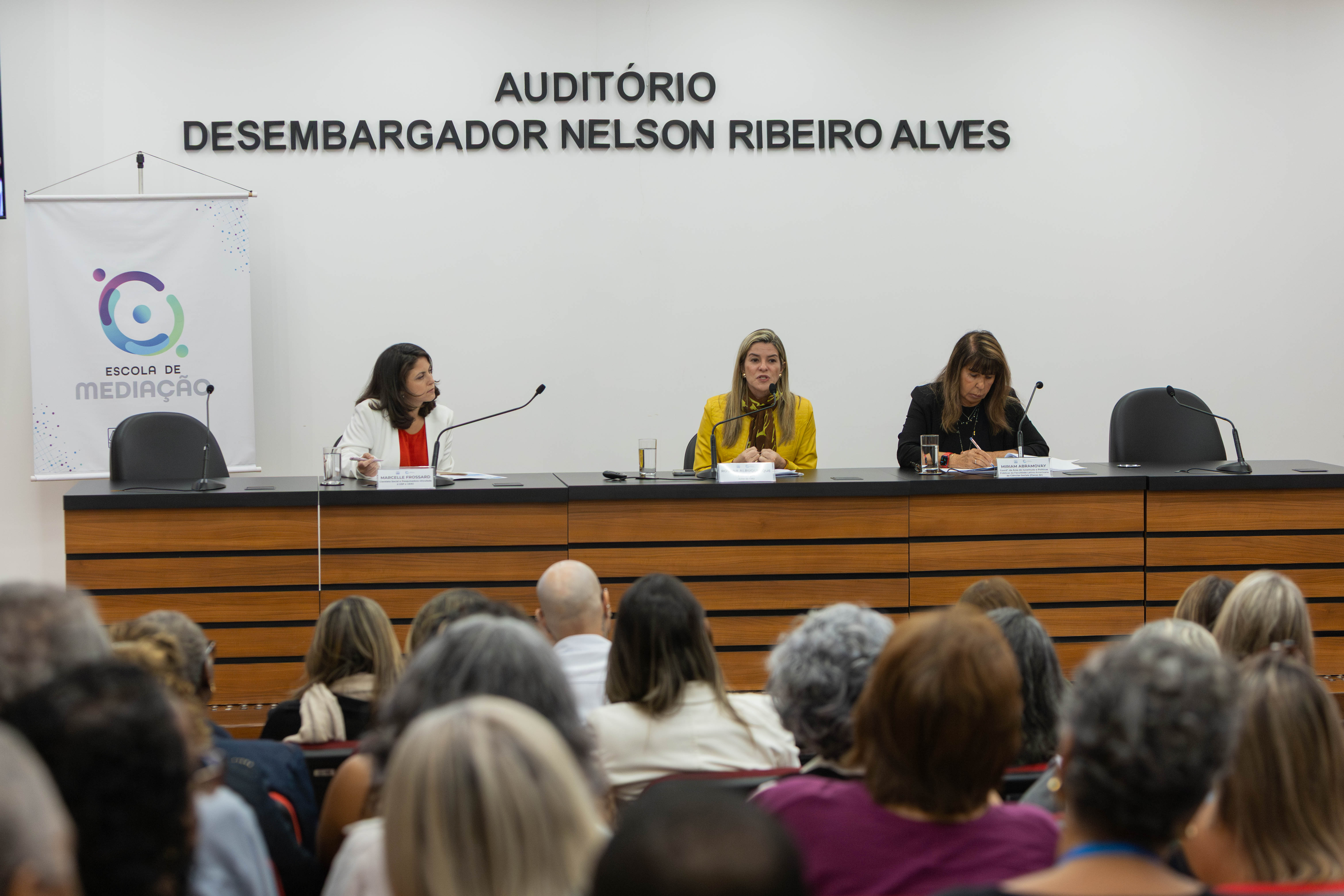 A imagem mostra uma palestra realizada no Auditório Desembargador Nelson Ribeiro Alves. Em primeiro plano, o público acompanha atentamente a mesa de debates posicionada à frente do espaço. Na mesa principal, três mulheres participam do encontro. A participante ao centro, vestindo blazer amarelo, fala ao microfone enquanto as demais acompanham a apresentação e fazem anotações. Sobre a bancada há microfones, copos com água e documentos. À esquerda da imagem, um banner da Escola de Mediação identifica a instituição organizadora do evento. O ambiente possui paredes claras, acabamento em madeira e iluminação uniforme.